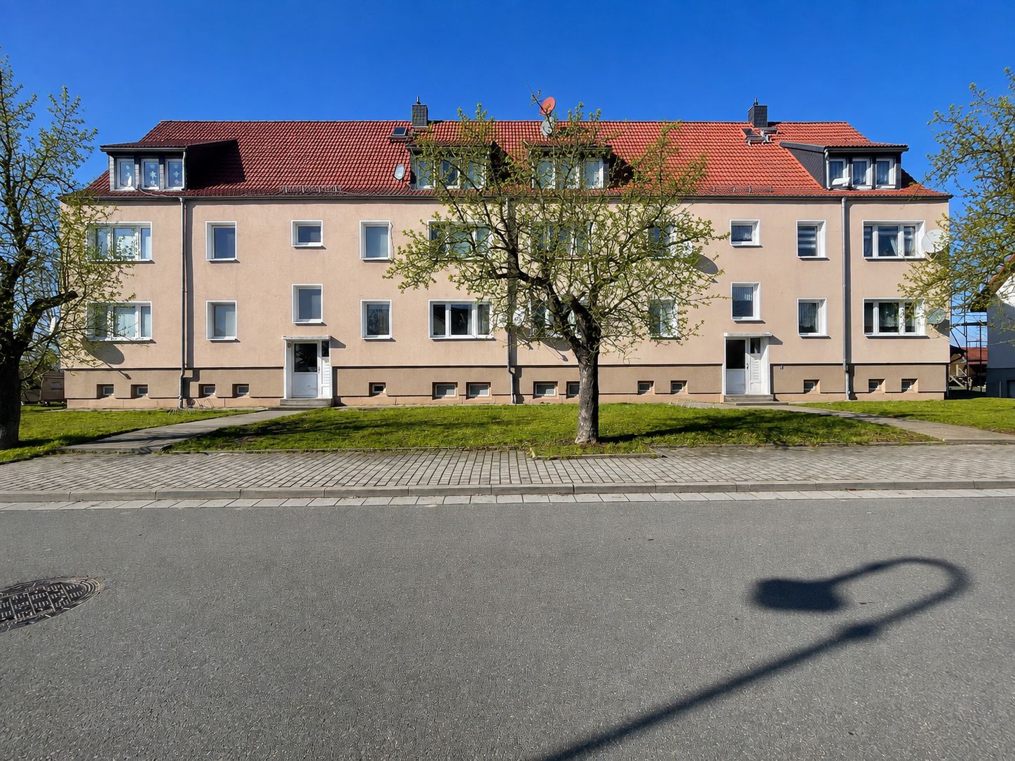 Mehrfamilienhaus mit rotem Dach, mehreren Fenstern und Eingängen, davor Rasen und Bäume.