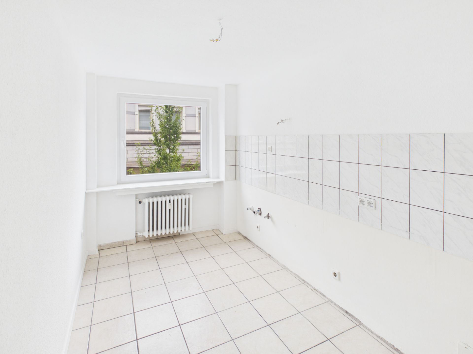 Leerer, gefliester Raum mit großem Fenster und Heizkörper, weiße Wände und Fliesen.