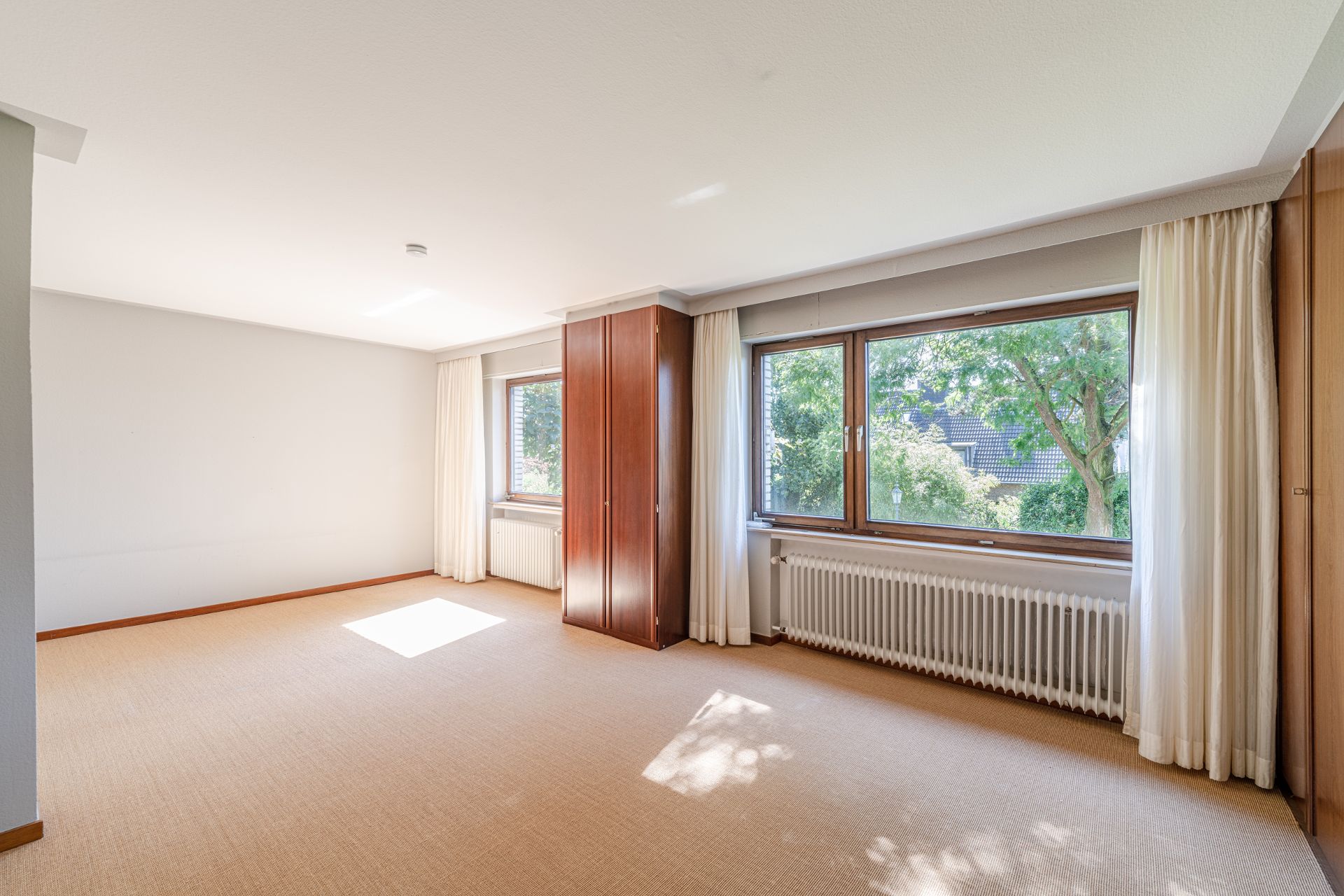 Helles, leeres Zimmer mit zwei großen Fenstern, Vorhängen und zwei Holzschränken.
