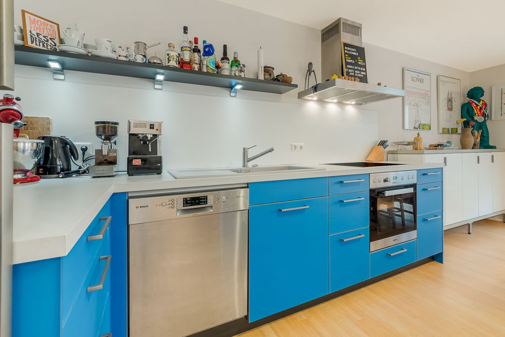 Moderne Küche mit blauen Unterschränken, Spülmaschine, Herd und offenen Regalen mit Küchenutensilien.