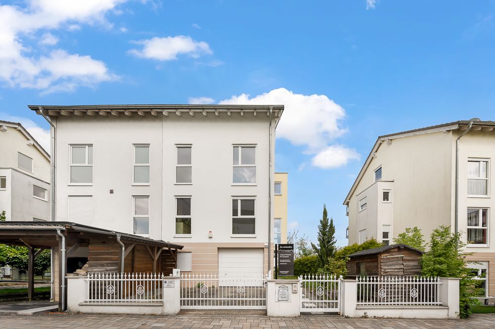 Modernes, weißes Wohnhaus mit drei Stockwerken, Vorgarten und Carport, umgeben von Zaun.
