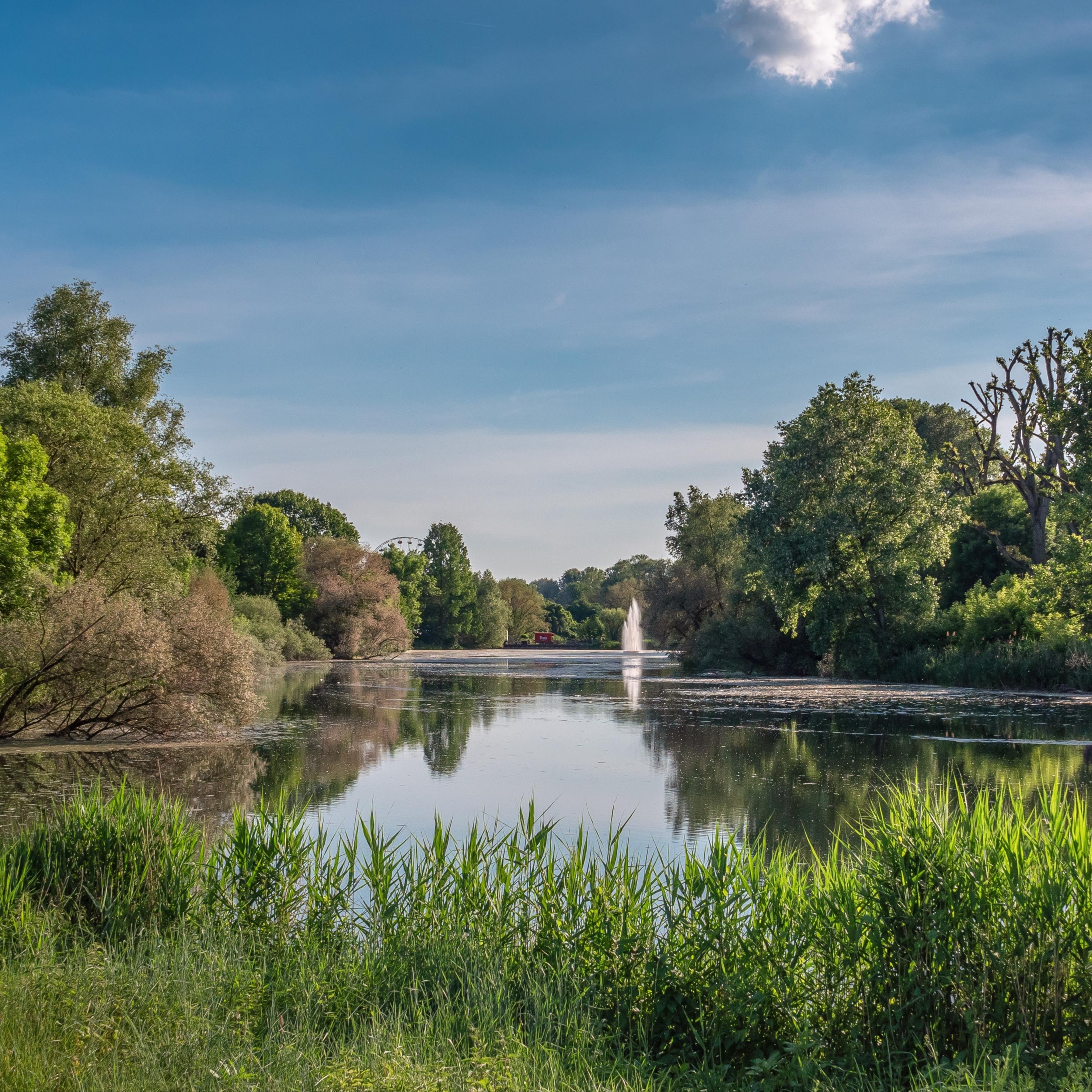 See mit Springbrunnen, umgeben von Bäumen und grüner Vegetation unter blauem Himmel.