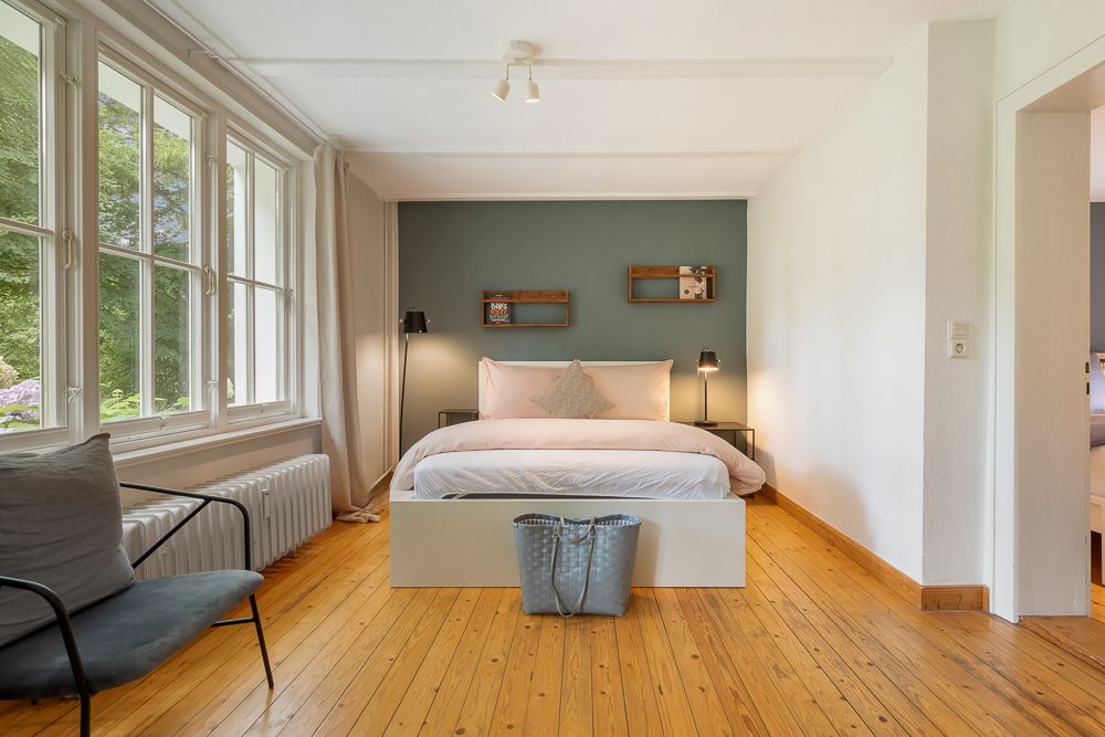 Helles Schlafzimmer mit großem Bett, Holzboden und großen Fenstern, daneben ein grauer Stuhl.