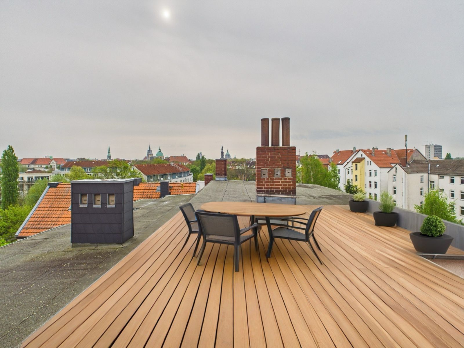 Dachterrasse mit Holzboden, Tisch, Stühlen und Blumentöpfen, umgeben von Wohnhäusern.
