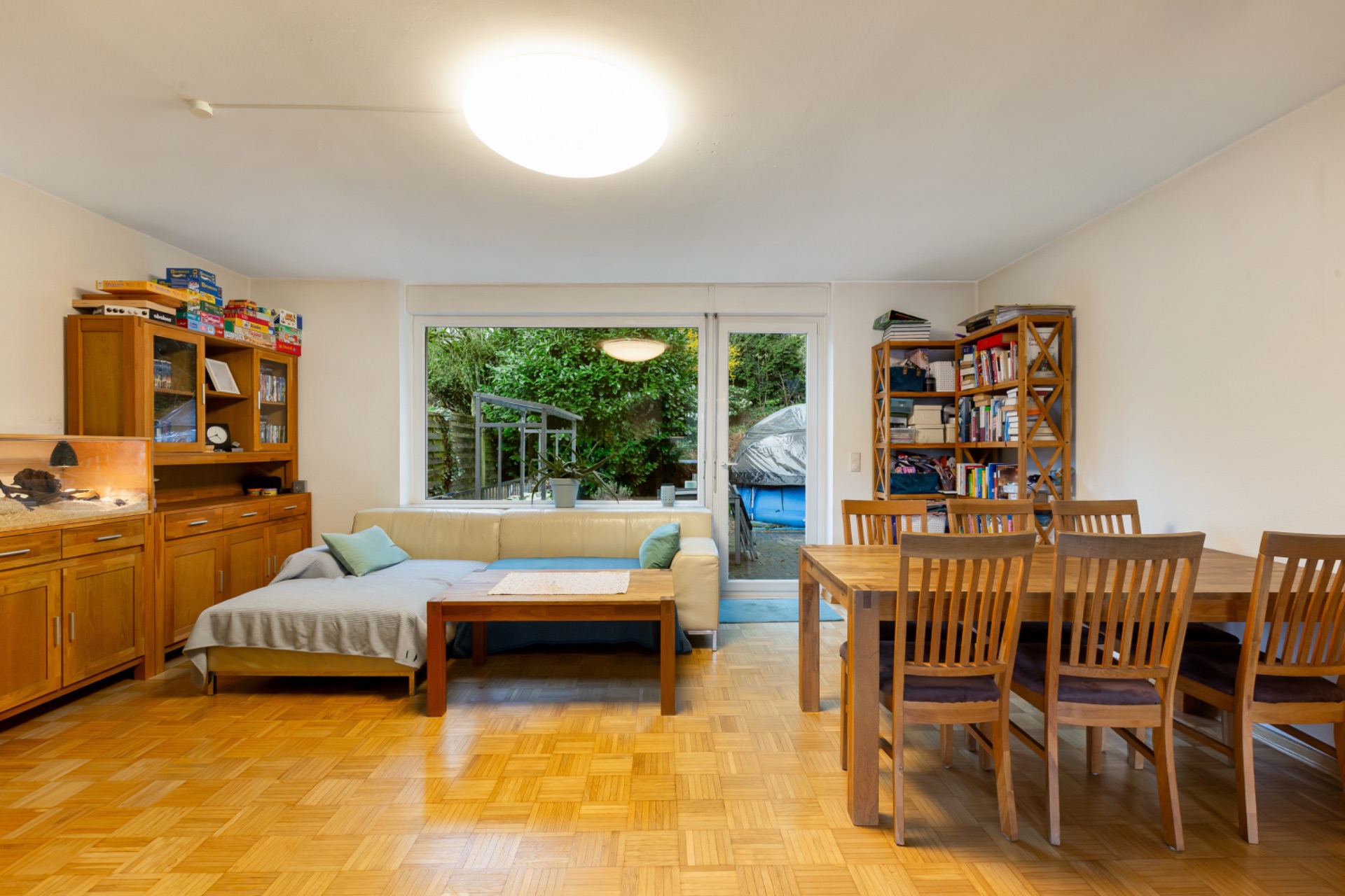 Wohnzimmer mit Holztisch, Stühlen, Sofa, Bücherregal und Blick auf den Garten durch große Fenster.