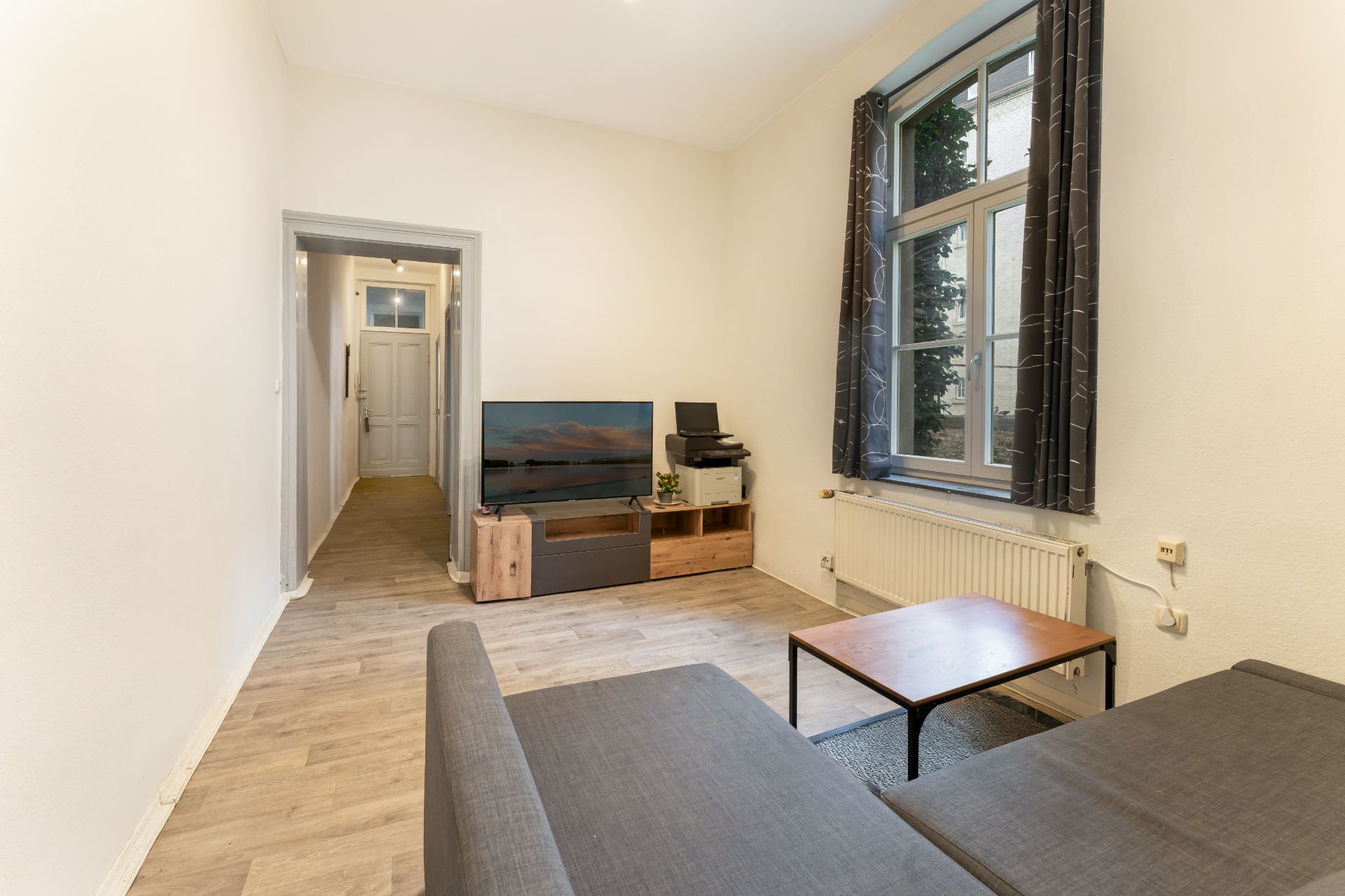 Wohnzimmer mit grauem Sofa, Holztisch, Fernseher, Fenster mit Vorhängen und Holzfußboden.