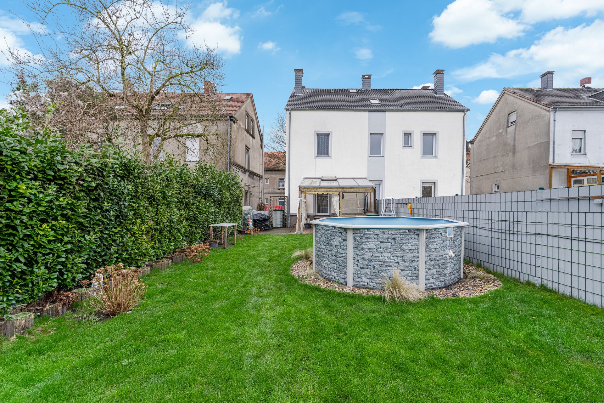 Grüner Garten mit rundem Aufstellpool hinter einem weißen Wohnhaus.