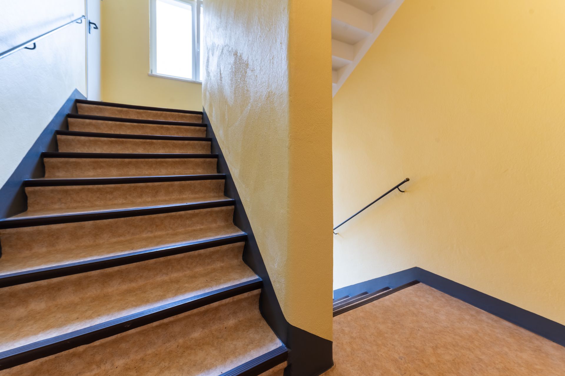 Innenansicht eines Treppenhauses mit gelben Wänden und Fenster am Treppenabsatz.