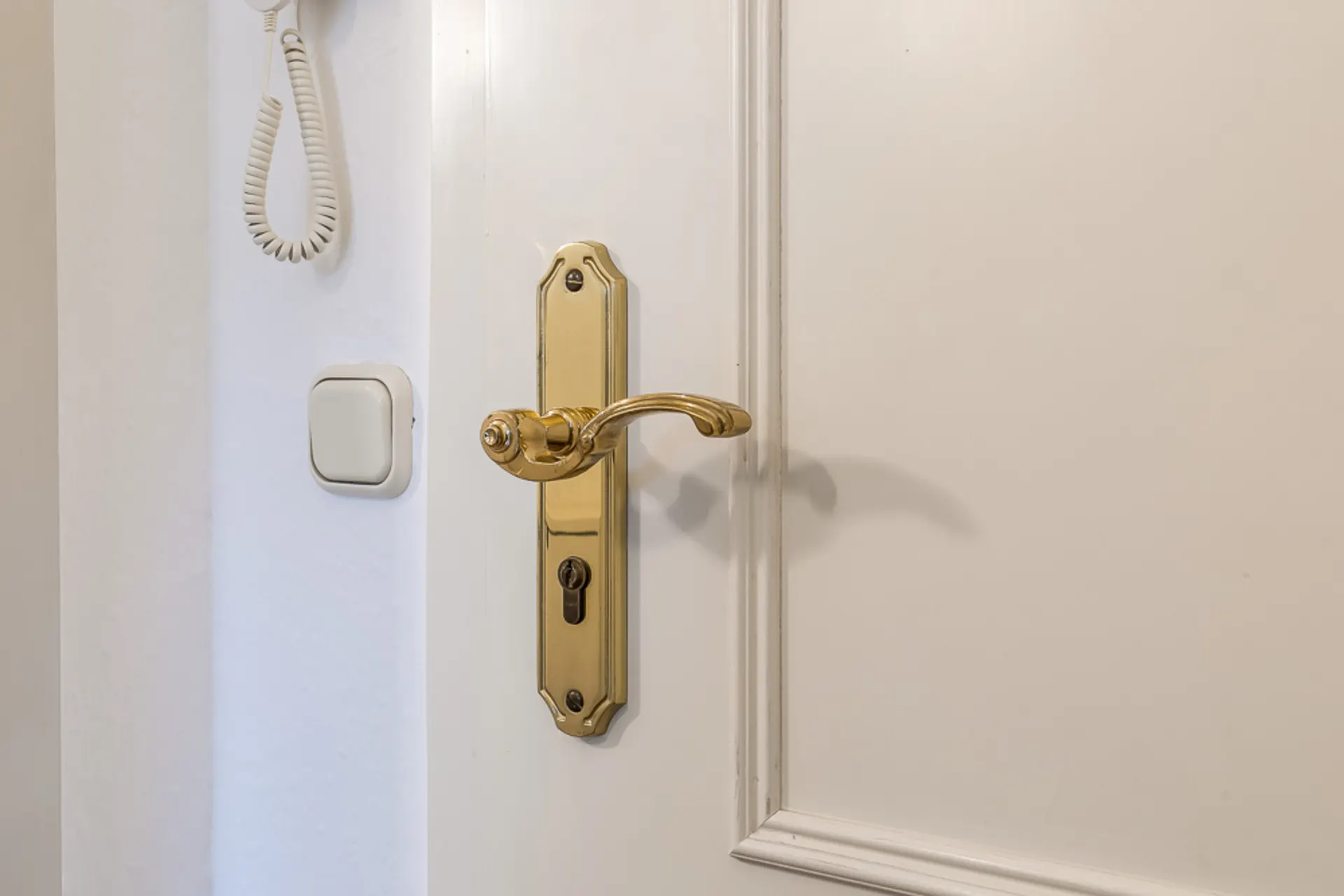 Nahaufnahme eines weißen Türblatts mit goldenem Griff und Lichtschalter an der Wand.