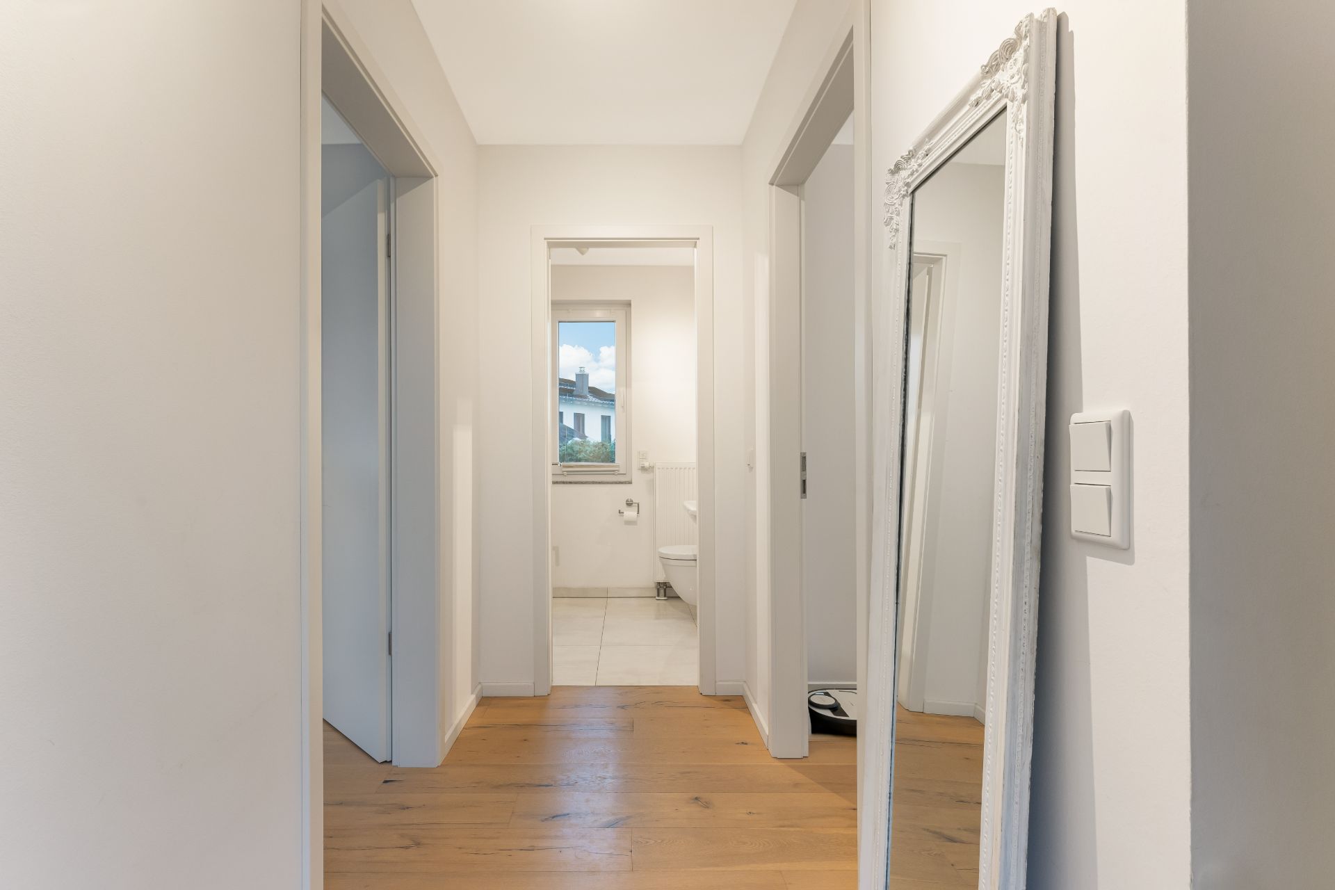 Heller Flur mit Holzboden, Wandspiegel und Blick in ein Badezimmer mit Fenster.