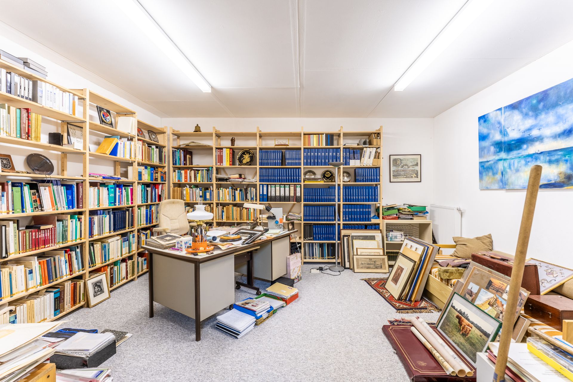 Arbeitszimmer mit Schreibtisch, vielen Büchern in Regalen und einigen Bildern an den Wänden und am Boden.