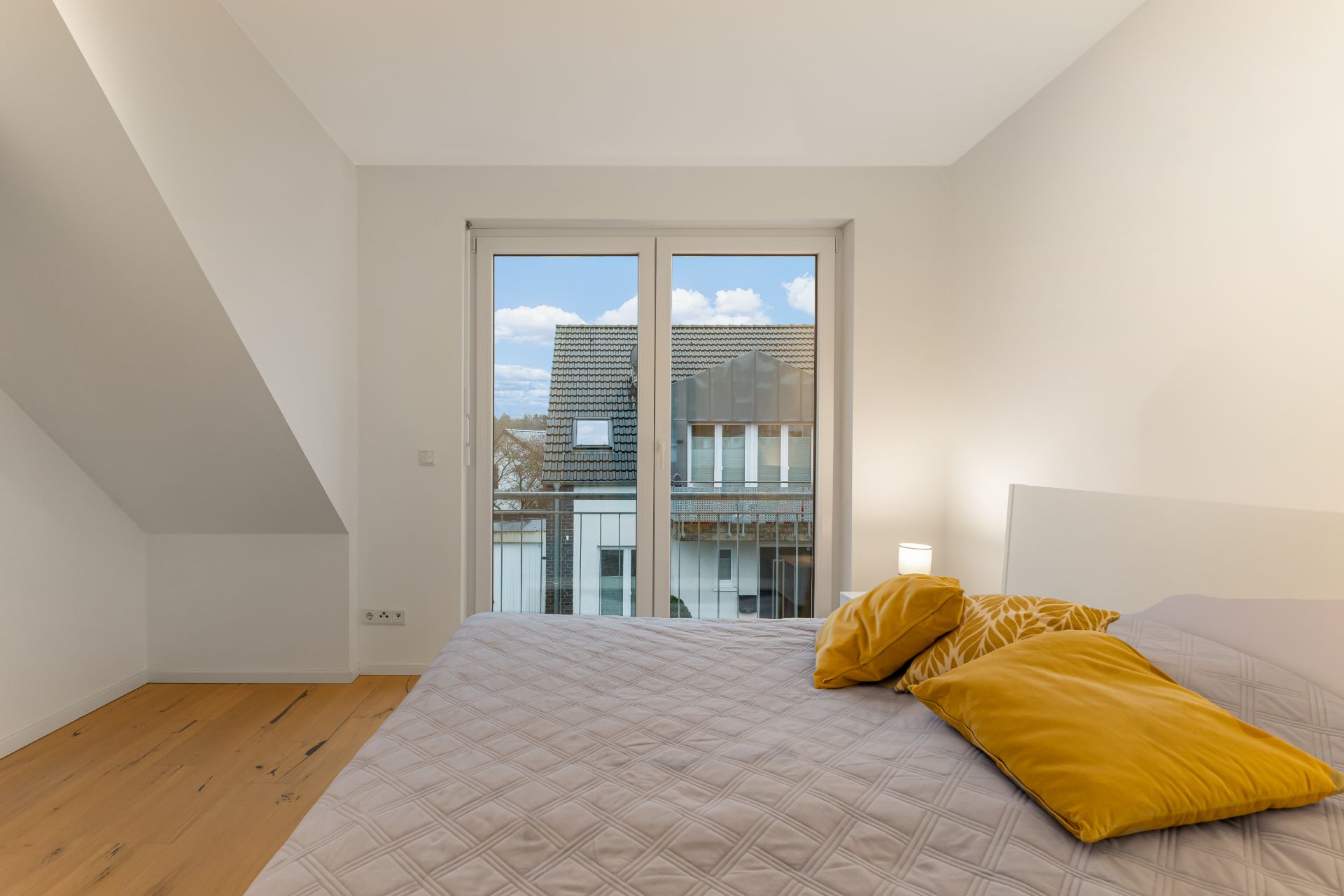 Helles Schlafzimmer mit großem Bett, gelben Kissen und Fenster mit Blick auf Nachbarhaus.