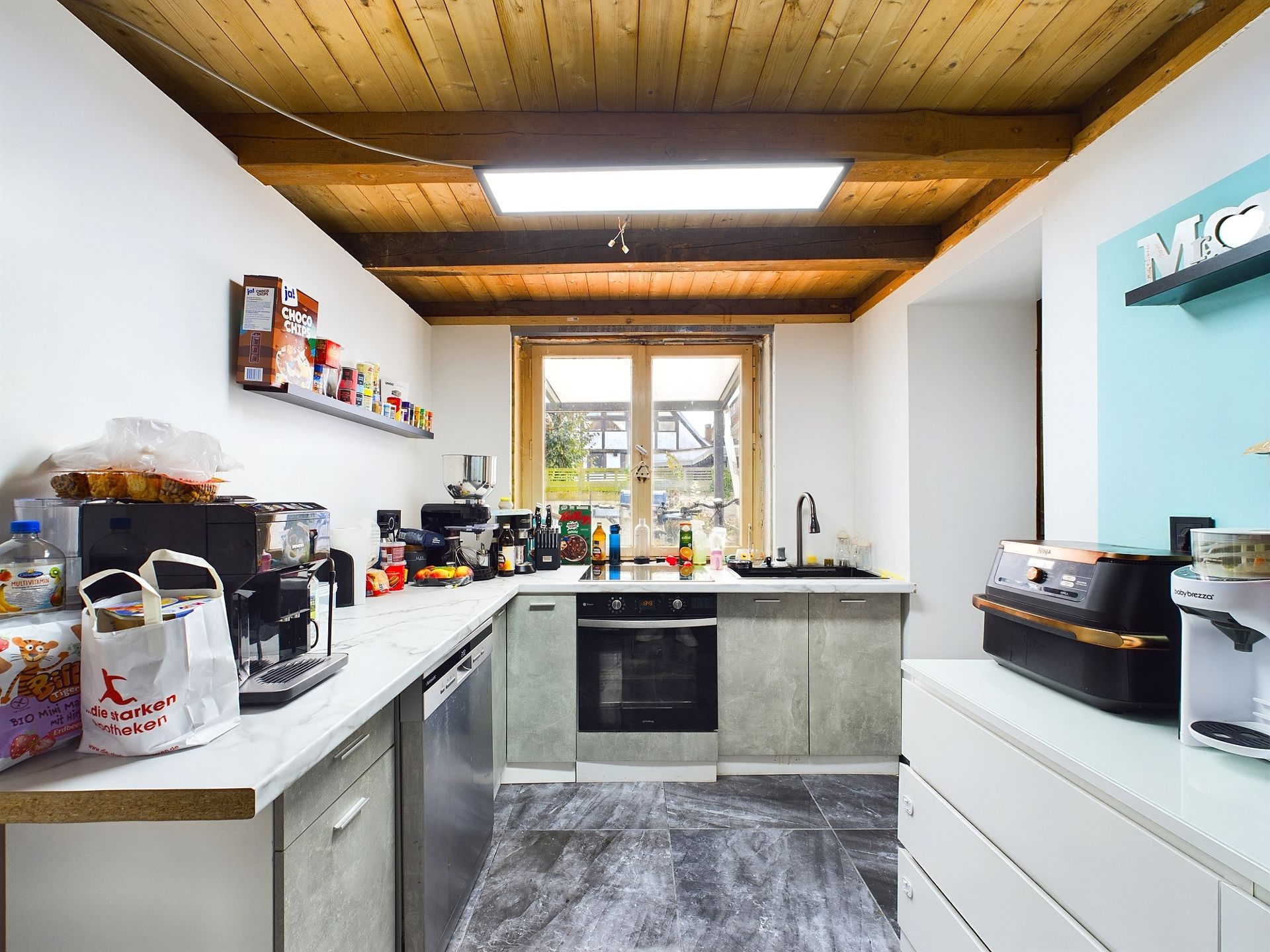 Moderne Küche mit Holzbalkendecke, großen Fenstern und verschiedenen Küchengeräten auf der Arbeitsfläche.