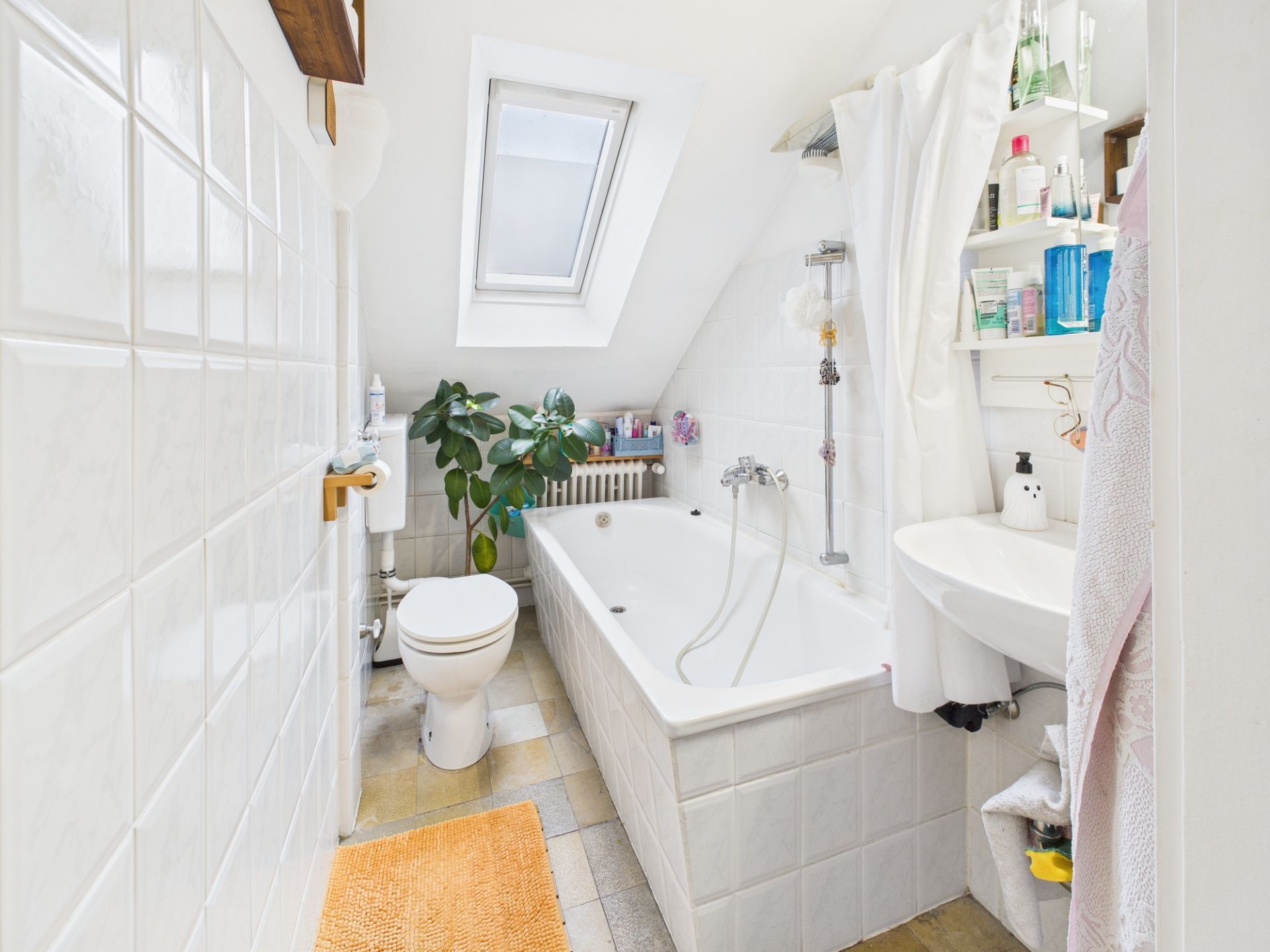Helles Badezimmer mit Badewanne, Toilette, Waschbecken, Regal, Pflanze und Dachfenster.