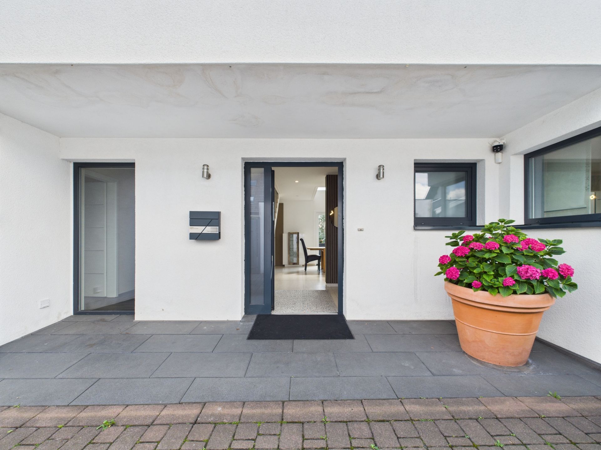 Eingang eines Hauses mit grauer Türmatte und großem Blumentopf mit rosa Blumen rechts.