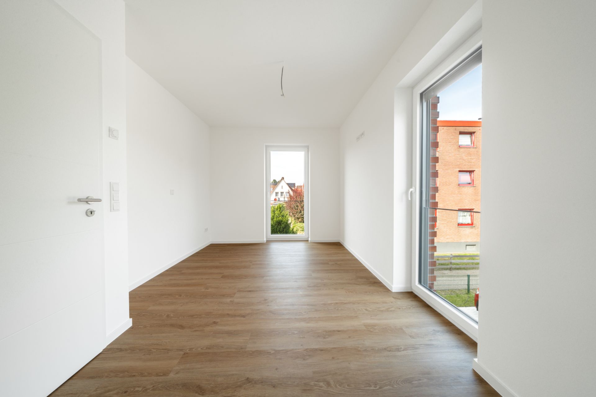 Helles leeres Zimmer mit Holzfußboden und großen Fenstern.