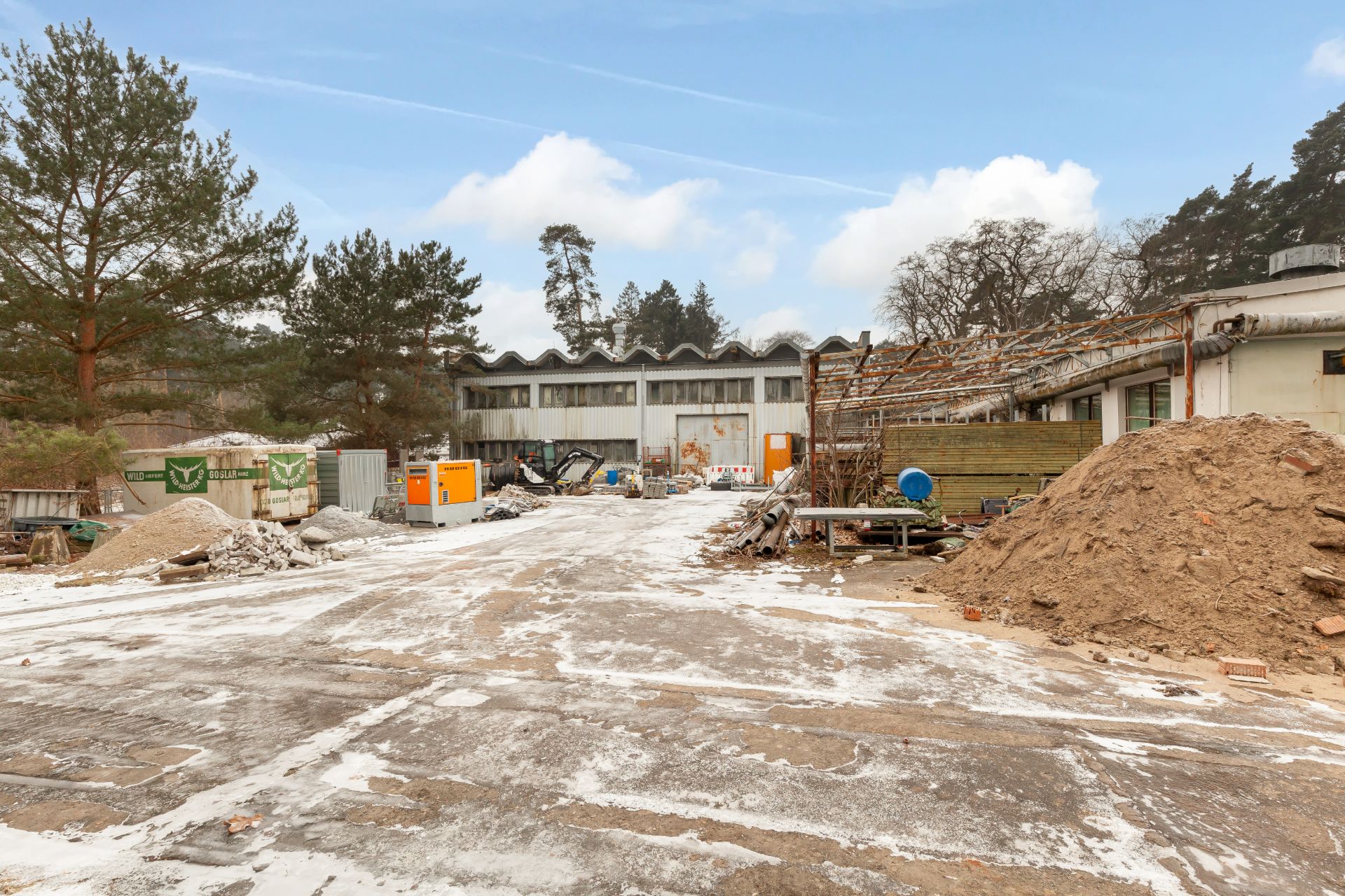 Verschneiter Hof mit Baucontainern, Baumaterial und Sandhaufen vor einem Gebäude.