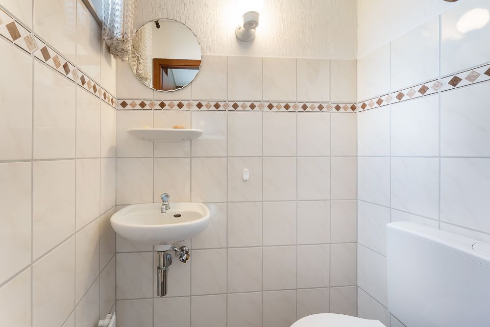 Kleines Badezimmer mit weiß gefliesten Wänden, Waschbecken, Spiegel, Ablage und WC.