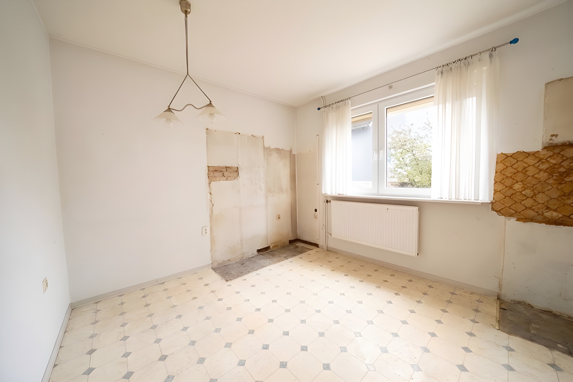 Leerer Raum mit hellen Wänden, Fenster, Heizkörper und abgenutztem Bodenbelag.
