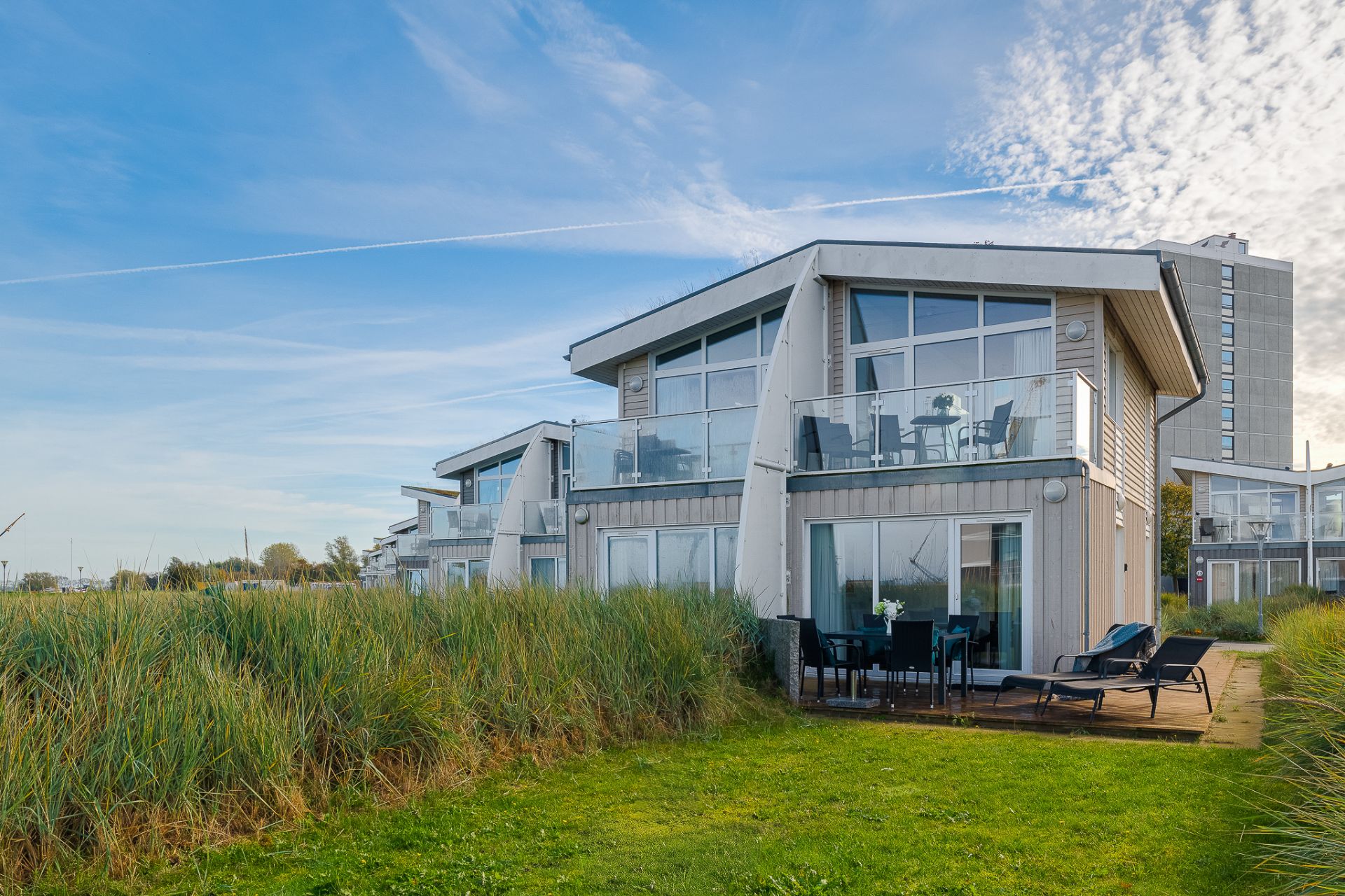 Modernes Wohnhaus mit großer Glasfront, Balkon und Terrasse, umgeben von Wiese und Pflanzen.