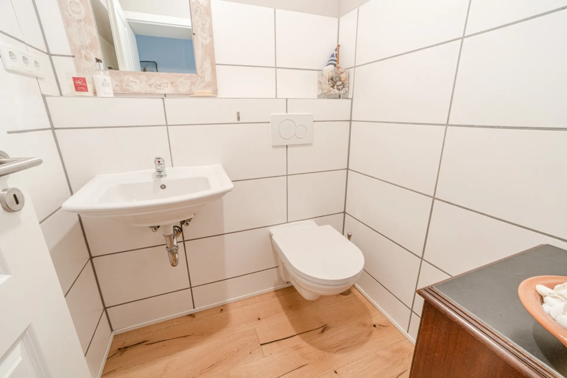Kleines Badezimmer mit weißem Waschbecken, Wandtoilette und Spiegel, Holzboden.