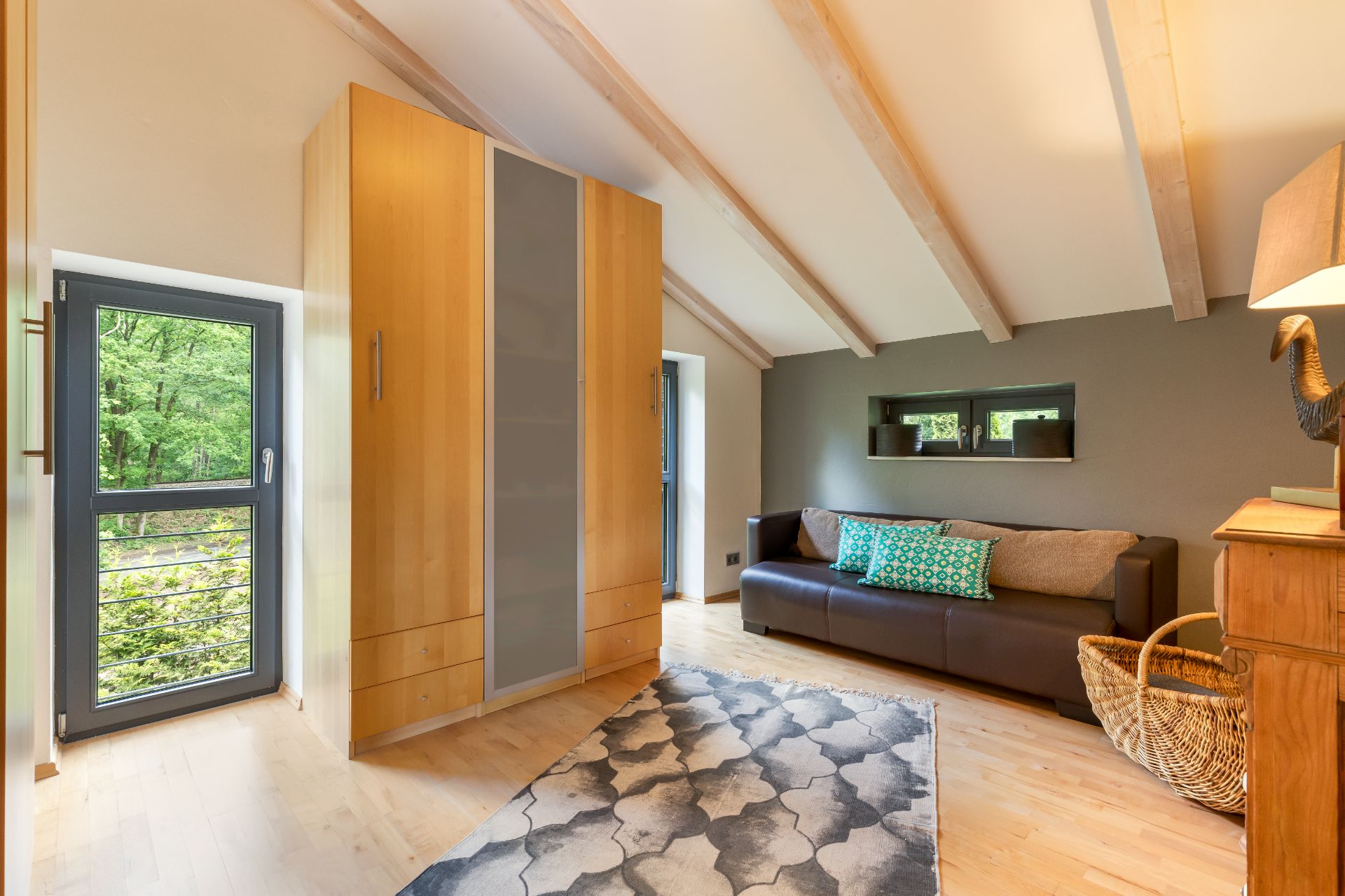 Wohnzimmer mit braunem Sofa, Holzschrank, Teppich und großen Fenstern mit Blick ins Grüne.