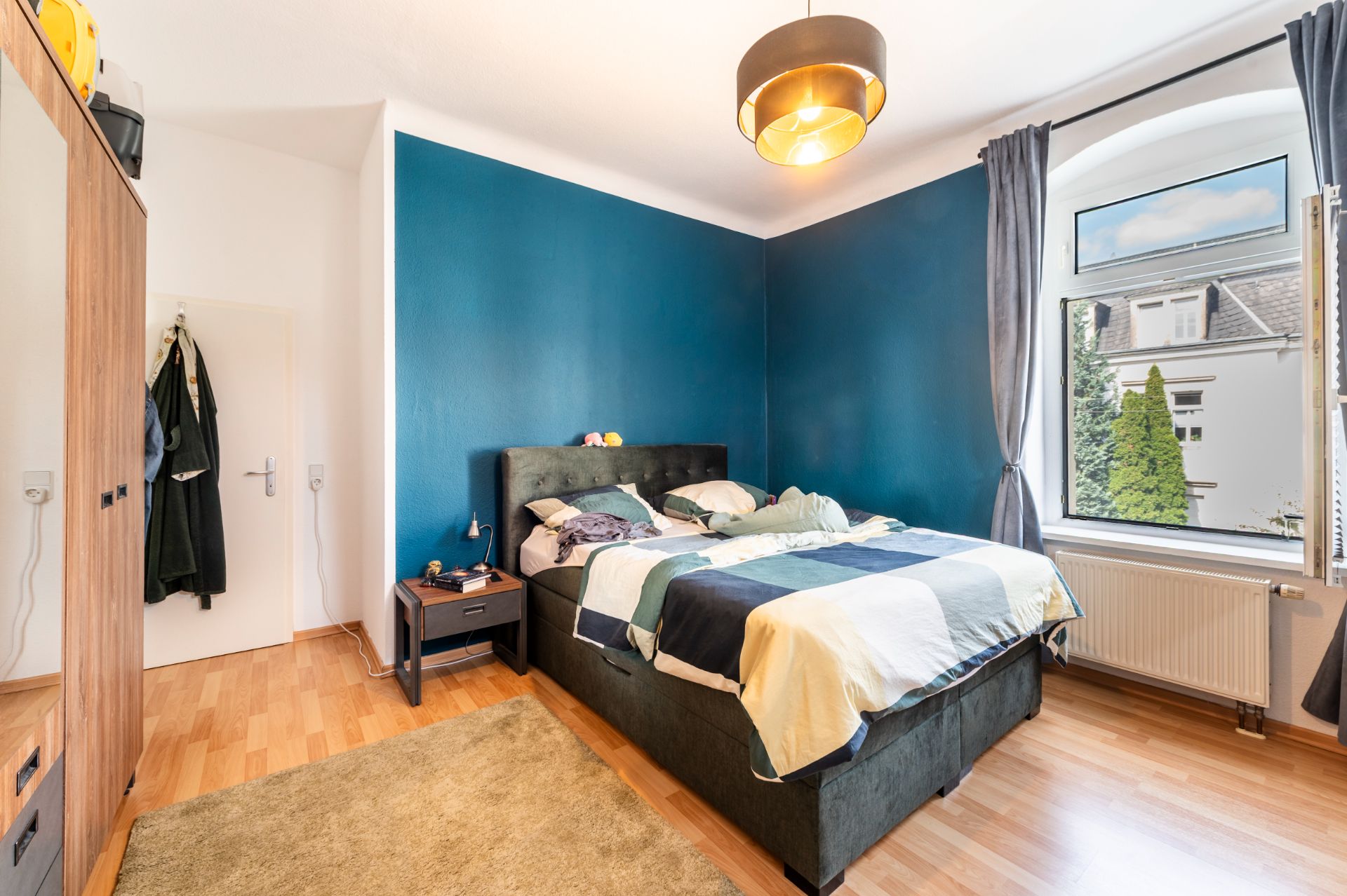 Schlafzimmer mit Doppelbett, blau gestrichener Wand, Fenster, Kleiderschrank und Nachttisch.