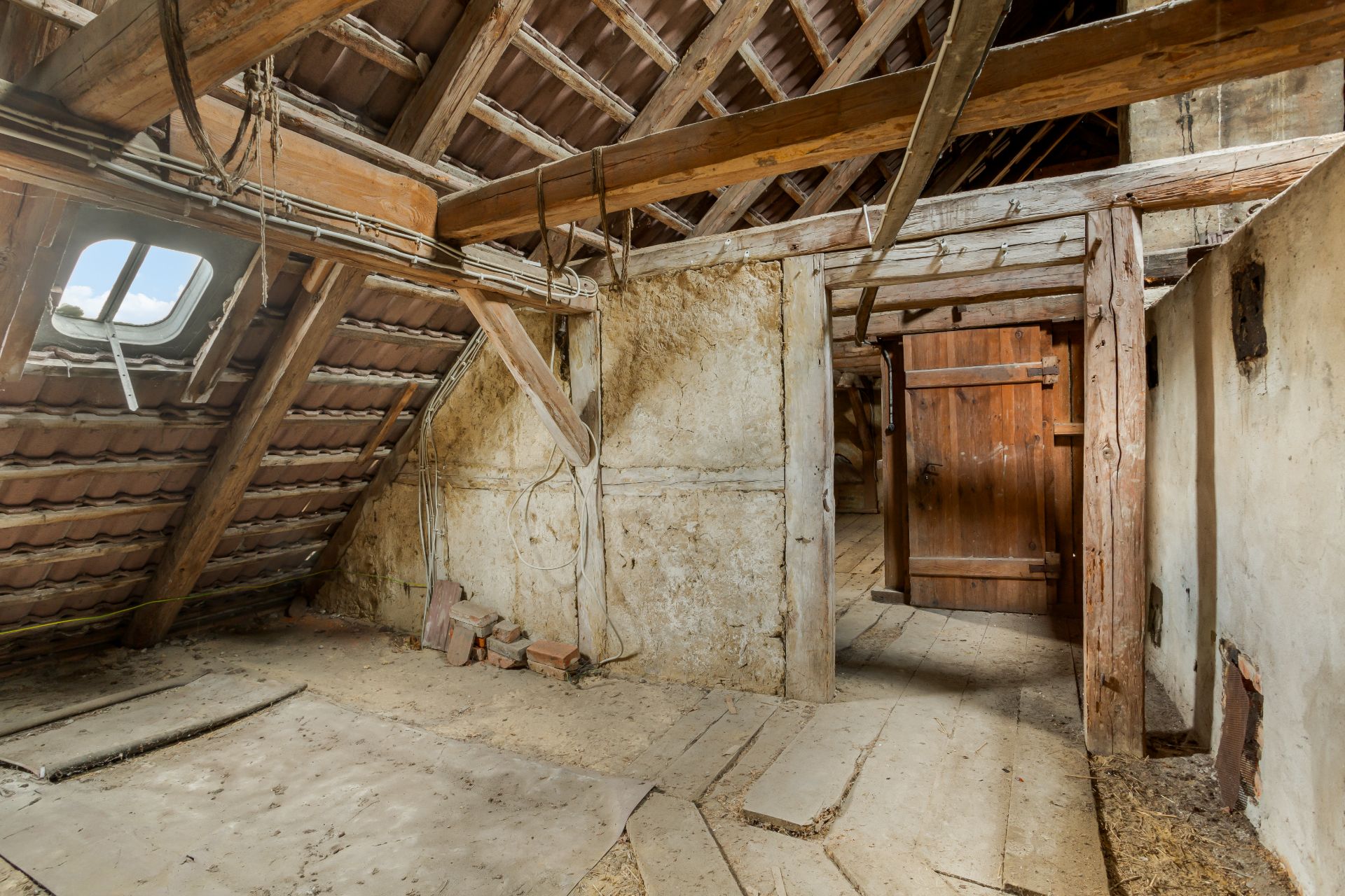 Alter Dachboden mit Holzbalken, Fenster und einer Holztür, teilweise renovierungsbedürftig.