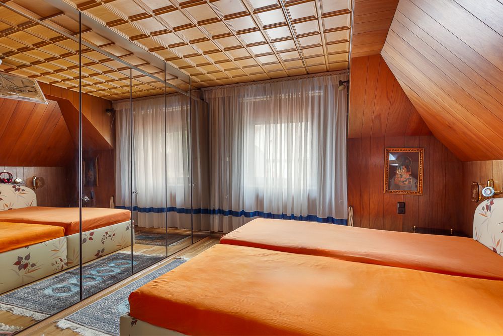 Schlafzimmer mit zwei Betten, orange Bettwäsche, Spiegelwand und großen Fenstern mit Vorhängen.