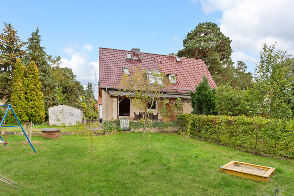 Zweistöckiges Haus mit rotem Dach und großem Garten mit Schaukel und Sandkasten.
