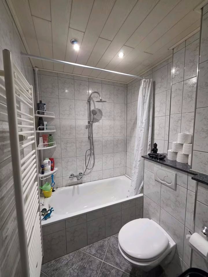 Kleines Badezimmer mit Badewanne, Dusche, WC, Heizkörper und Regal mit Toilettenpapier.