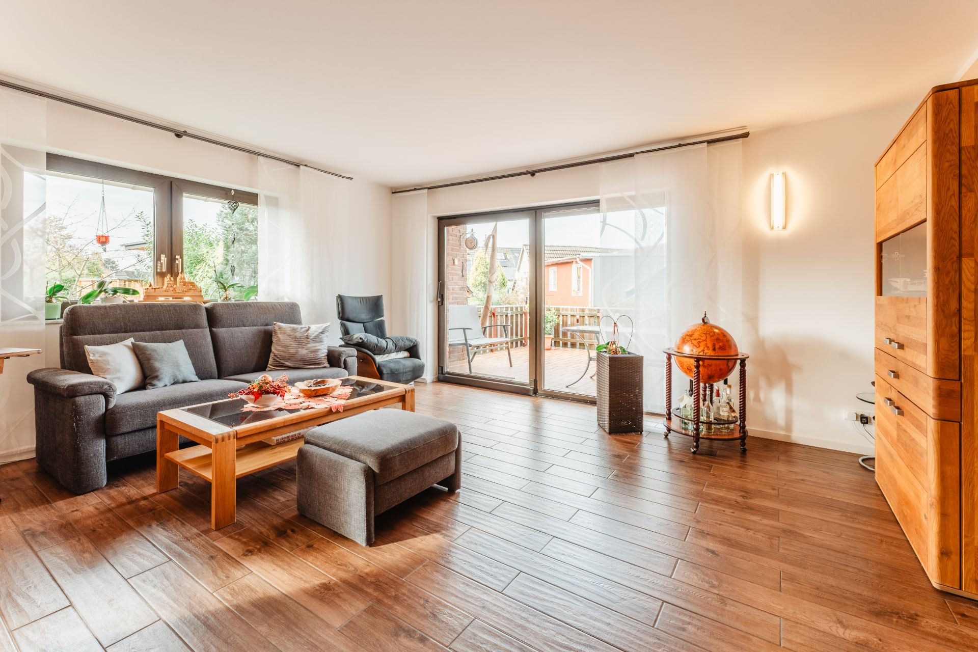 Helles Wohnzimmer mit grauem Sofa, Sessel, Glastisch und Holzfußboden, Zugang zur Terrasse.