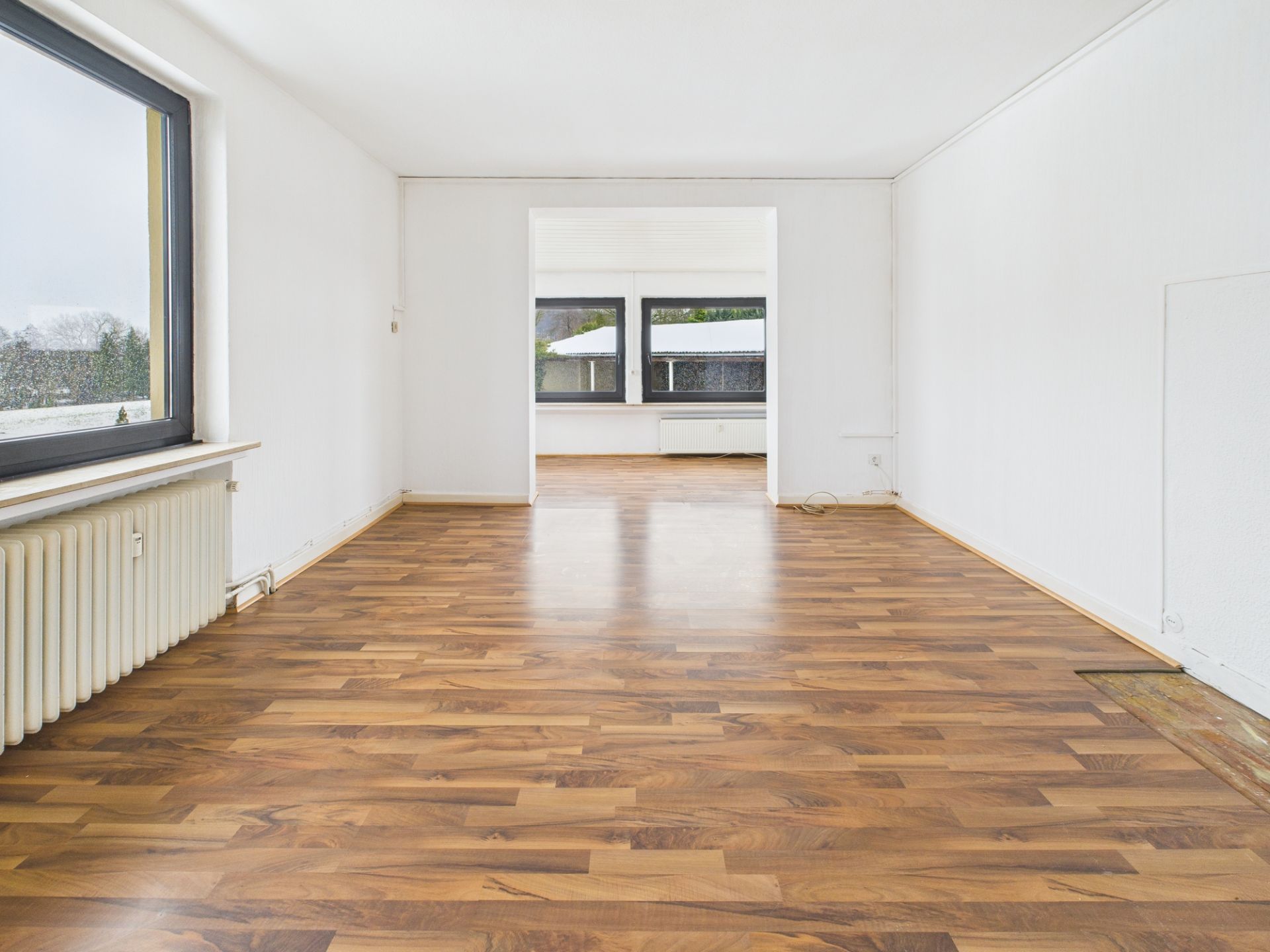 Leerer Raum mit Holzfußboden, weißen Wänden und großen Fenstern.