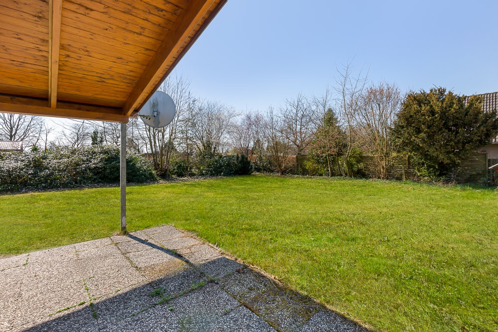 Überdachte Terrasse mit Blick auf einen großen, grünen Garten und einige Bäume im Hintergrund.