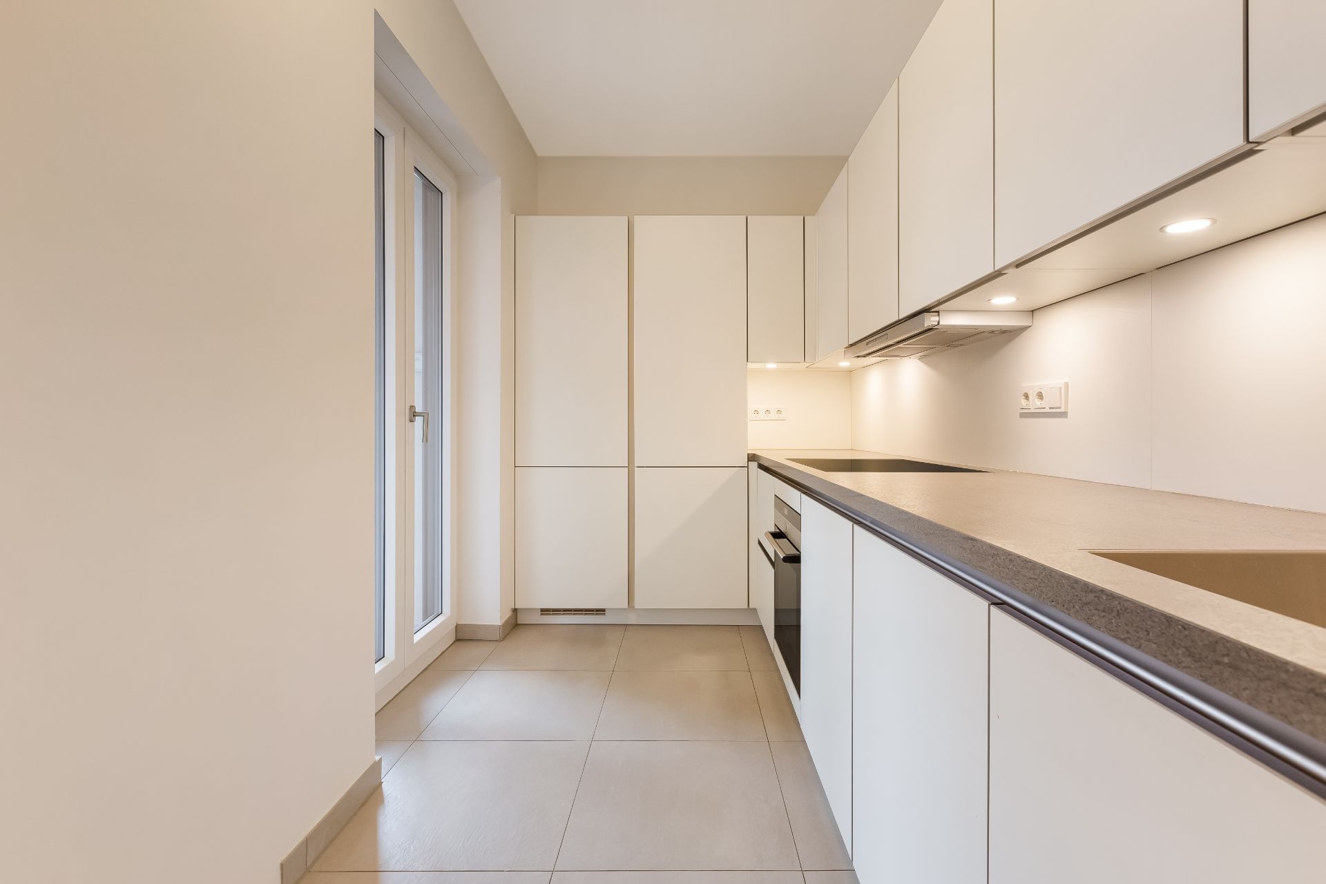 Moderne, helle Einbauküche mit weißen Schränken, grauer Arbeitsplatte und Fliesenboden.