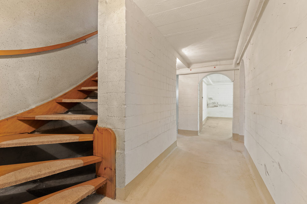 Heller Kellerflur mit weißen Wänden und einer offenen Holztreppe links.