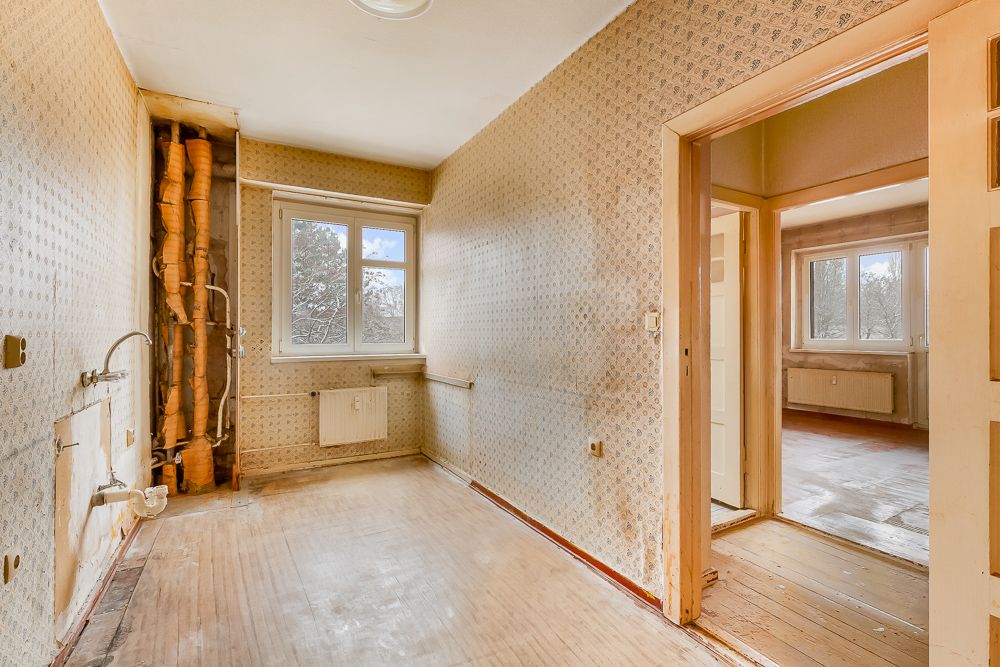Leerer, renovierungsbedürftiger Raum mit sichtbaren Rohren und zwei Fenstern.