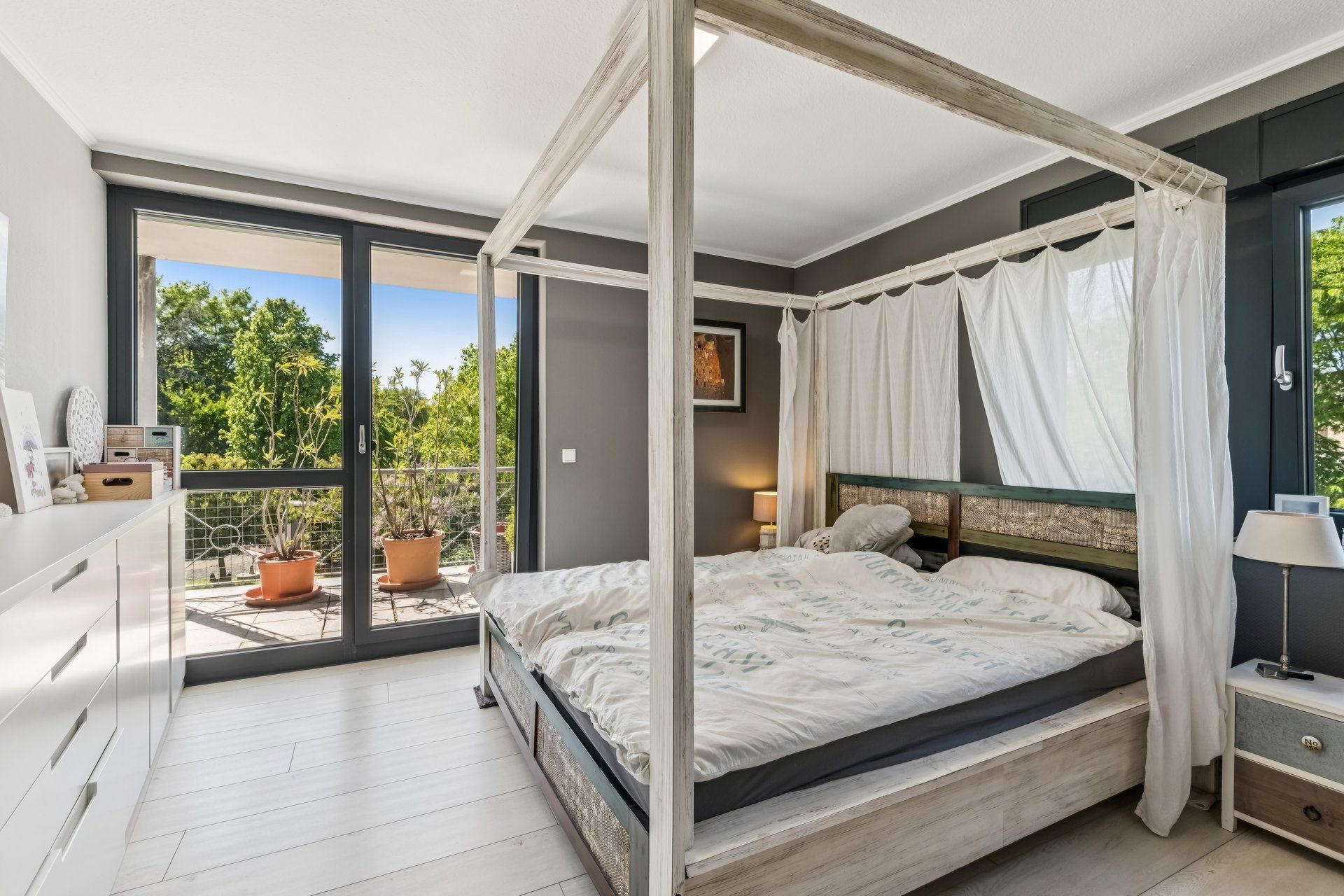 Helles Schlafzimmer mit Himmelbett, großer Fensterfront und Blick auf einen Balkon mit Pflanzen.