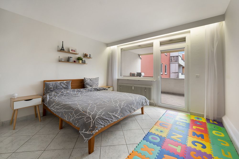 Schlafzimmer mit Doppelbett, Nachttisch, buntem Spielteppich und Zugang zu einem Balkon.
