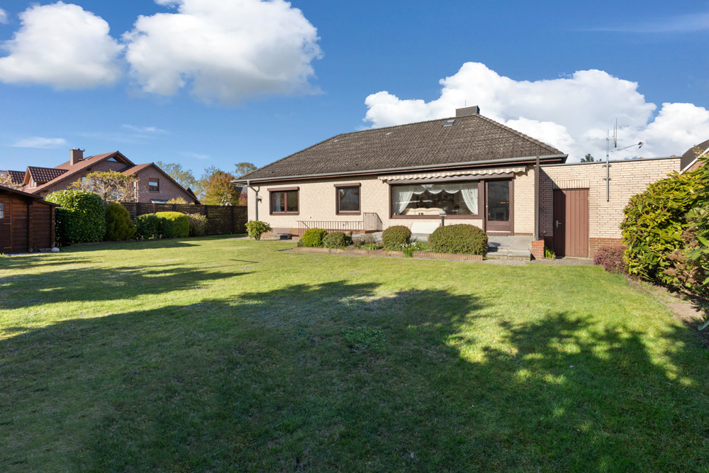 Einfamilienhaus mit Terrasse und großem Garten, umgeben von Büschen und Rasen.
