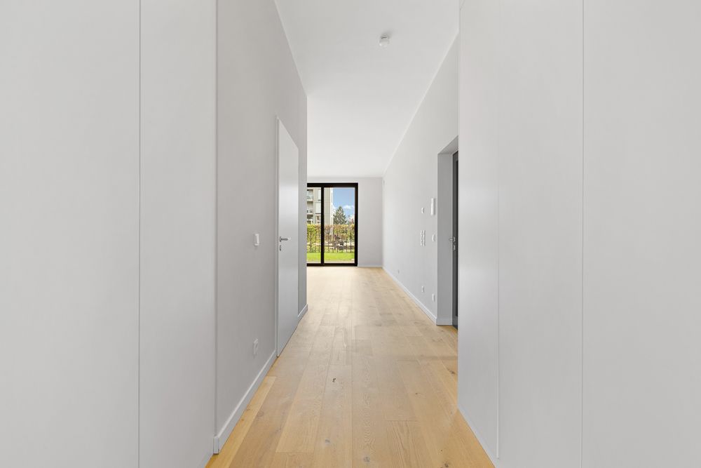 Heller Flur mit Holzboden und weißen Wänden, am Ende eine Glastür mit Blick ins Grüne.