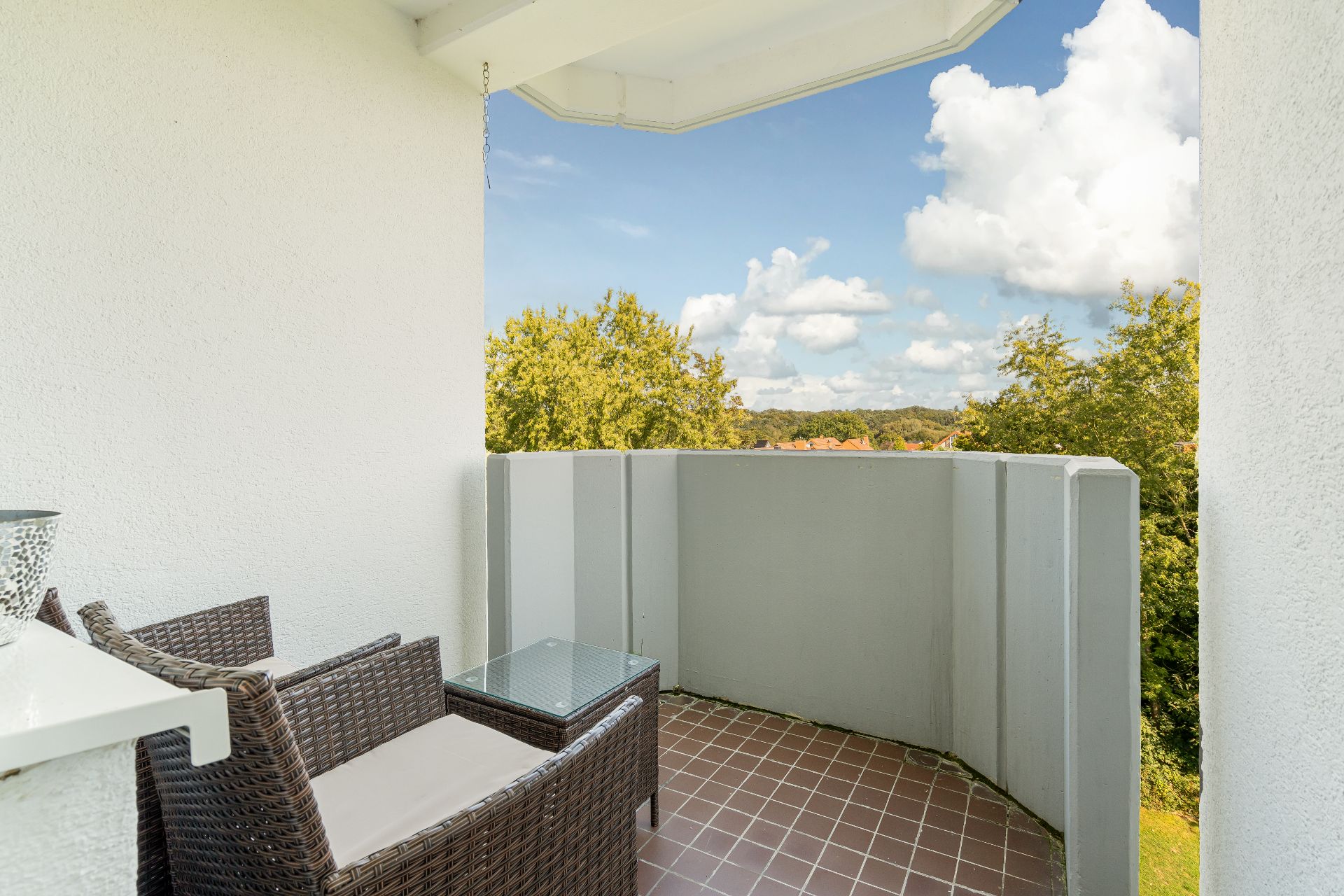 Kleiner Balkon mit zwei Rattan-Stühlen, Glastisch und Ausblick auf Bäume.