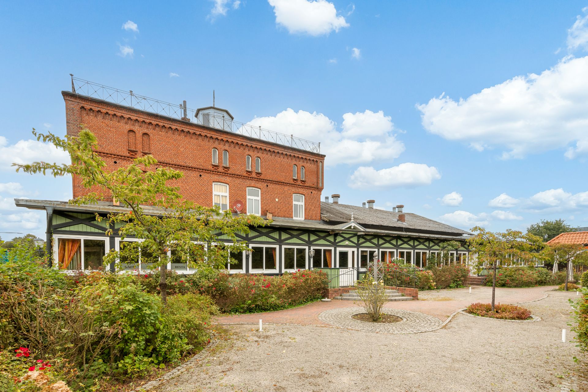 Backsteinwohnhaus mit Wintergarten, umgeben von Garten und gepflastertem Weg unter blauem Himmel.