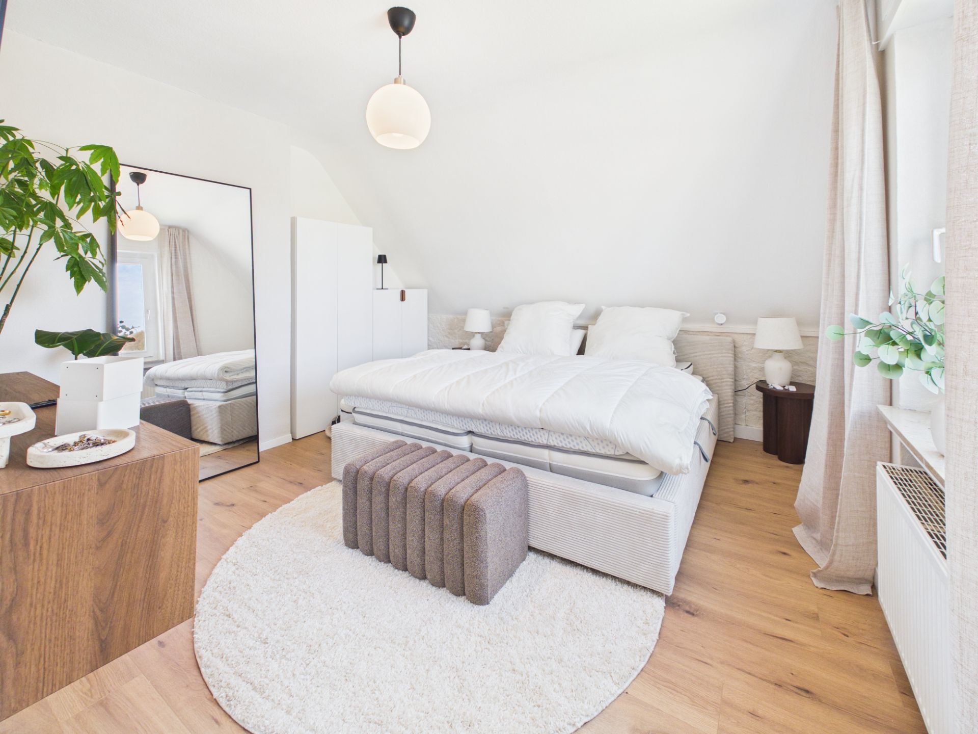 Helles Schlafzimmer mit Doppelbett, großem Spiegel, Holzkommode und Pflanzen auf hellem Holzboden.