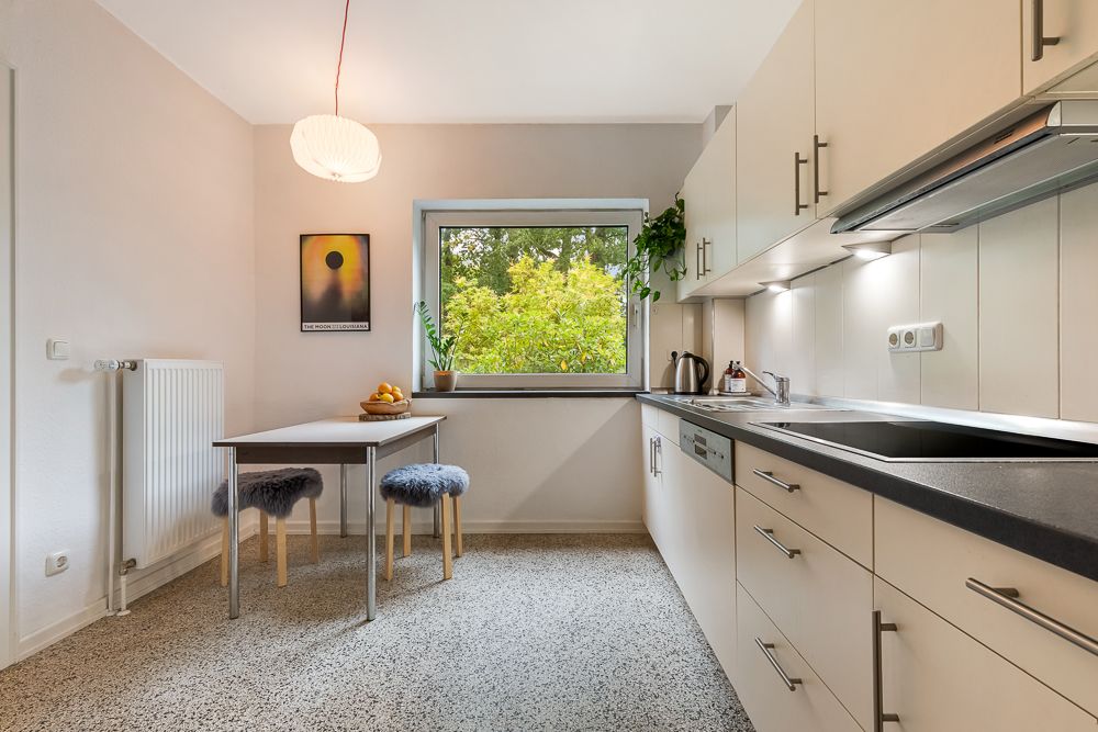 Helle Küche mit großem Fenster, Esstisch, zwei Hockern und Einbauküche.