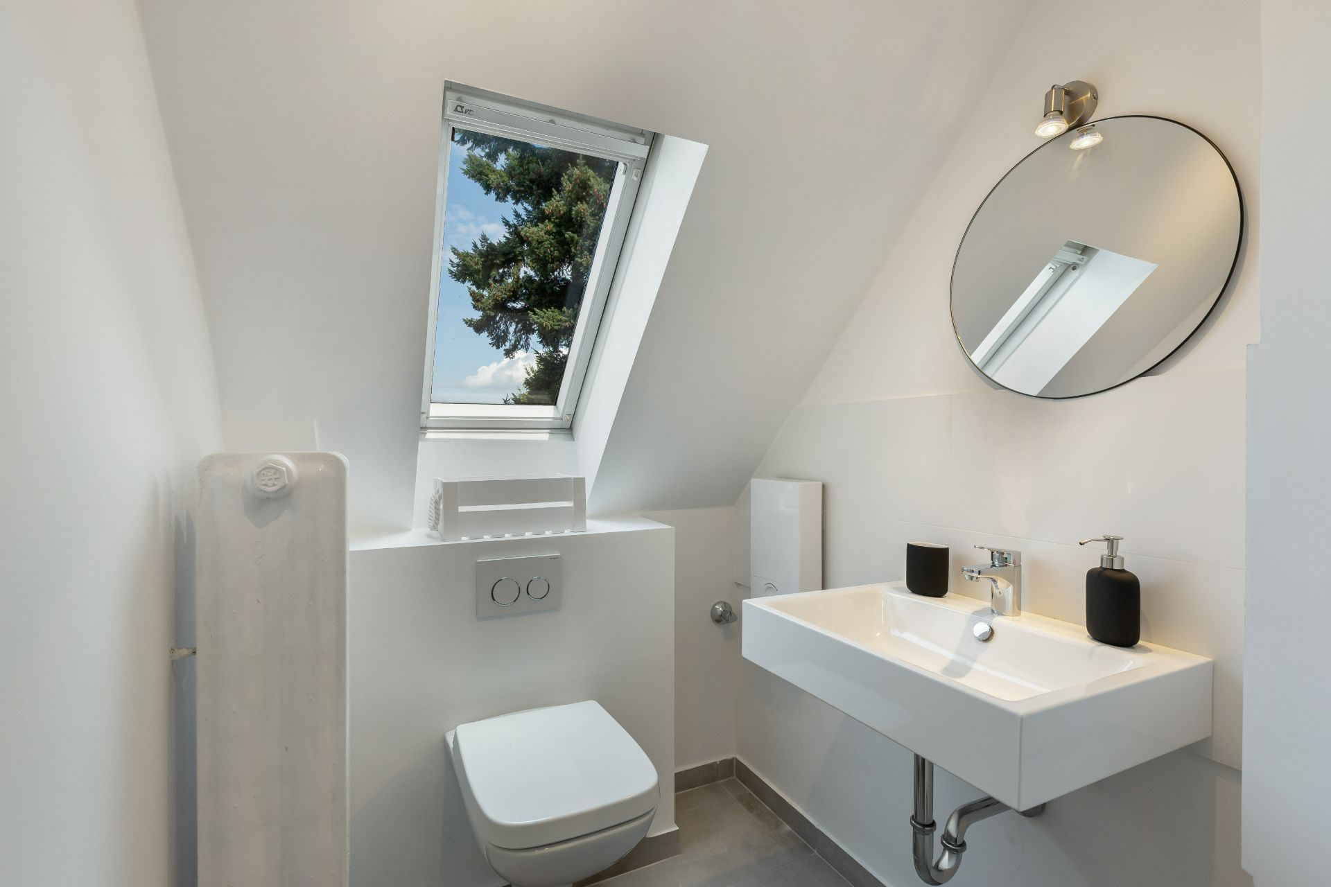 Modernes Badezimmer mit Dachfenster, Wandtoilette, Waschbecken und rundem Spiegel.