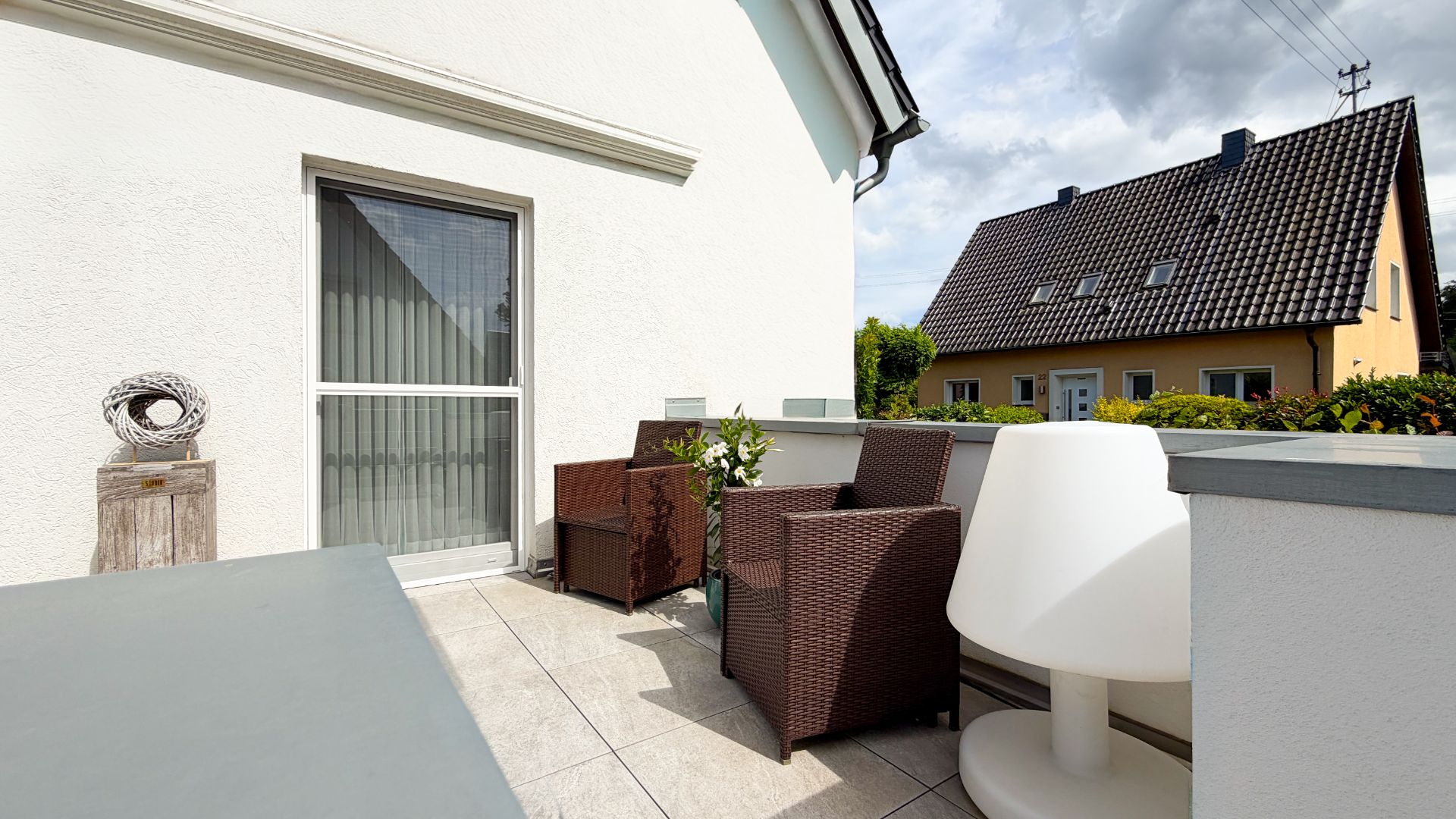 Helle Terrasse mit Sitzgruppe, Lampe und Blick auf Nachbarhaus, neben weißer Hauswand.
