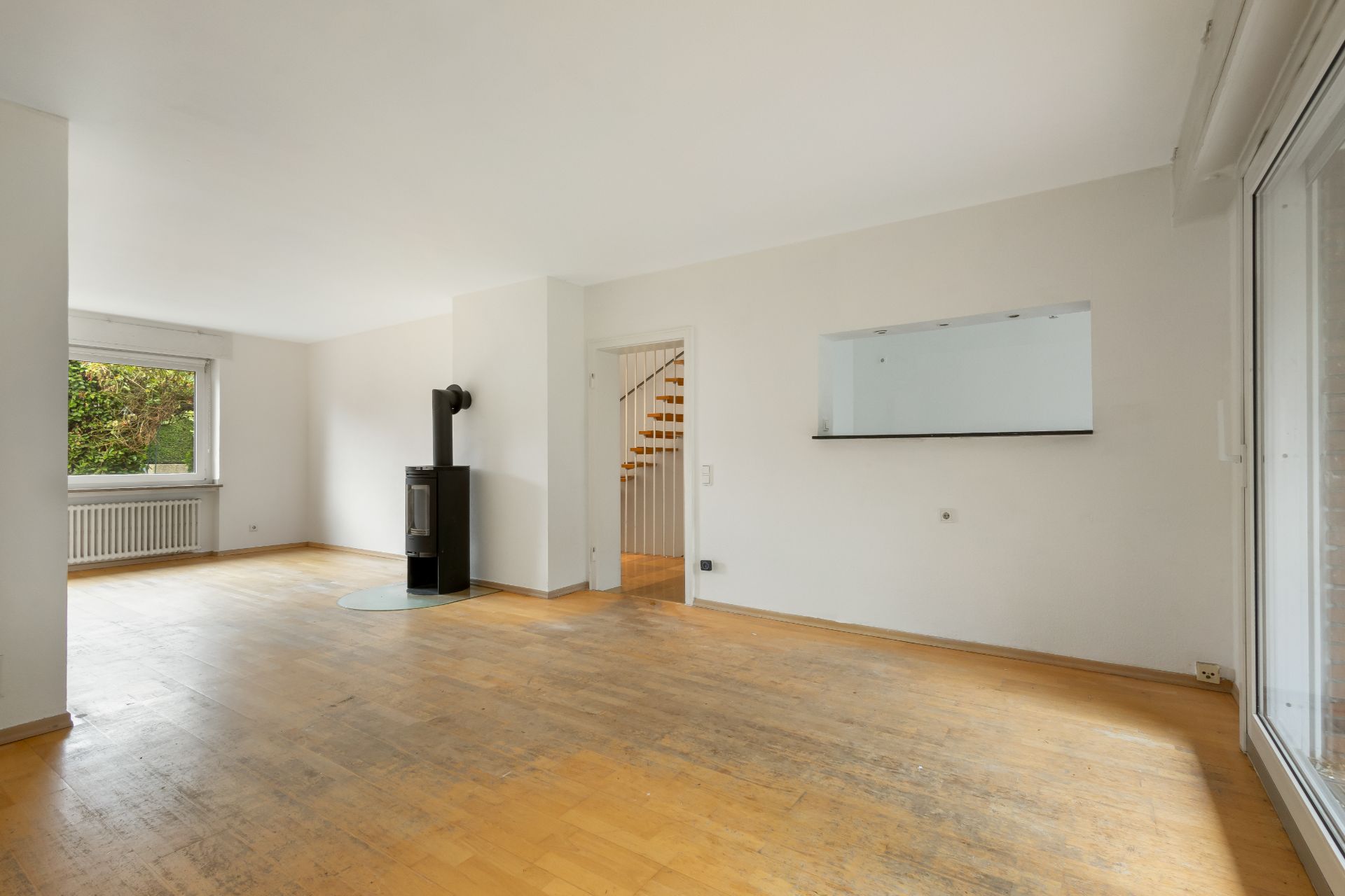 Helles, leeres Wohnzimmer mit Holzfußboden, Kaminofen und großem Fenster.