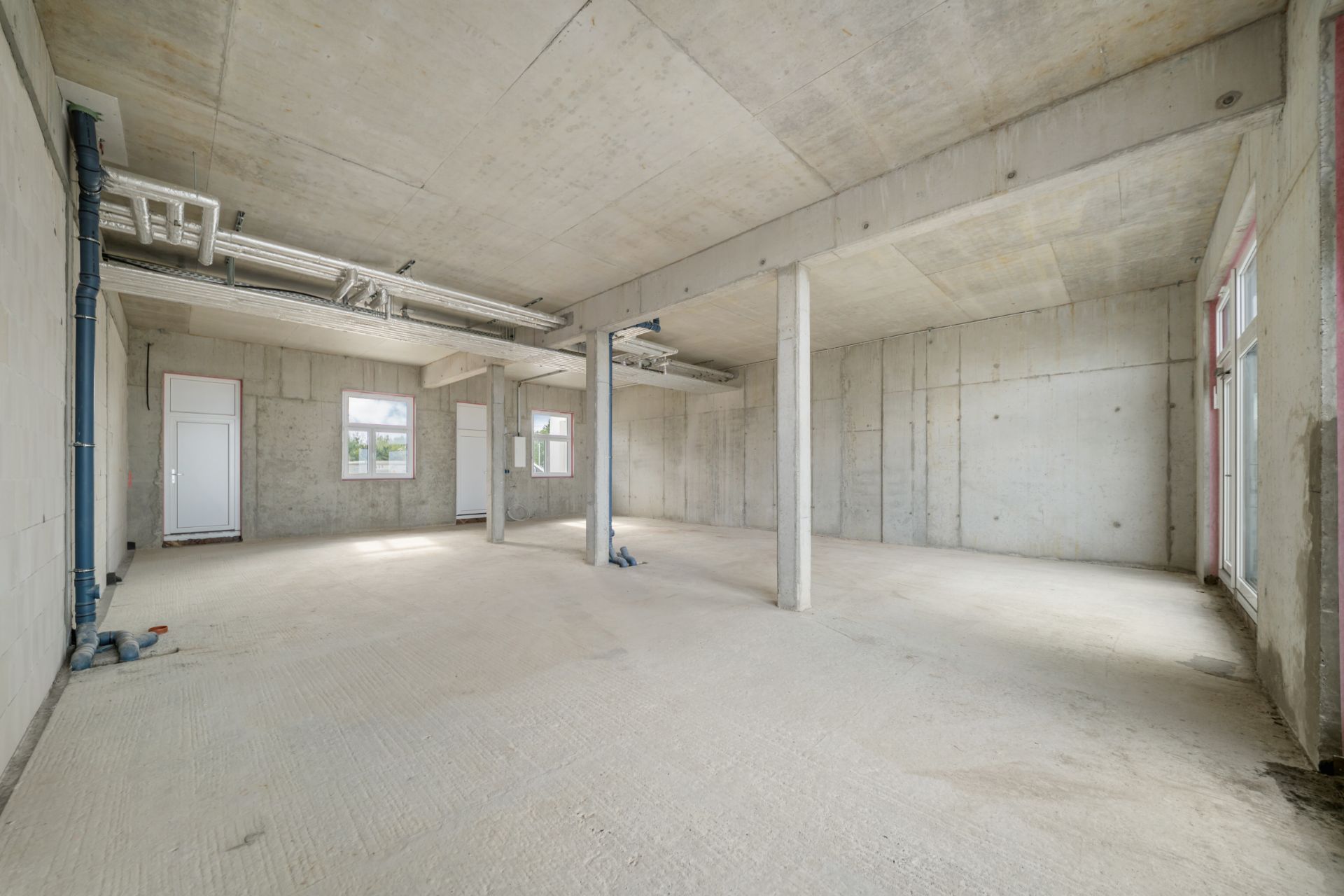Leerer Raum mit Betonwänden, Decke und Boden, mehreren Fenstern und sichtbaren Rohrleitungen.