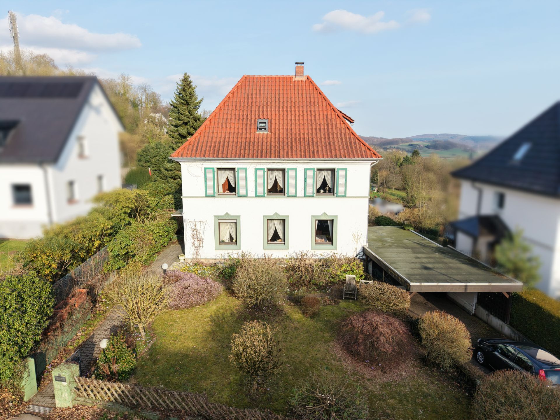 Zweistöckiges Haus mit rotem Dach, großem Garten und Carport auf der rechten Seite.